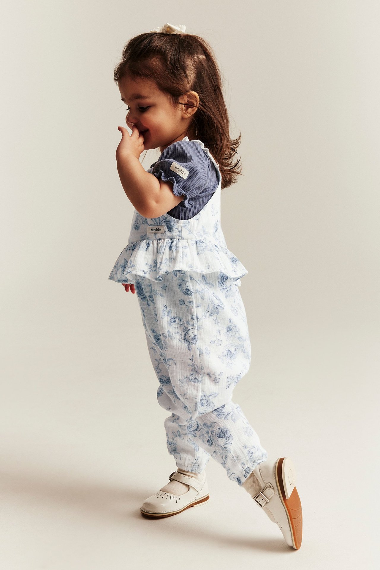 Side view: Kid wears off-white floral dungarees with frill over a blue T-shirt and white shoes.