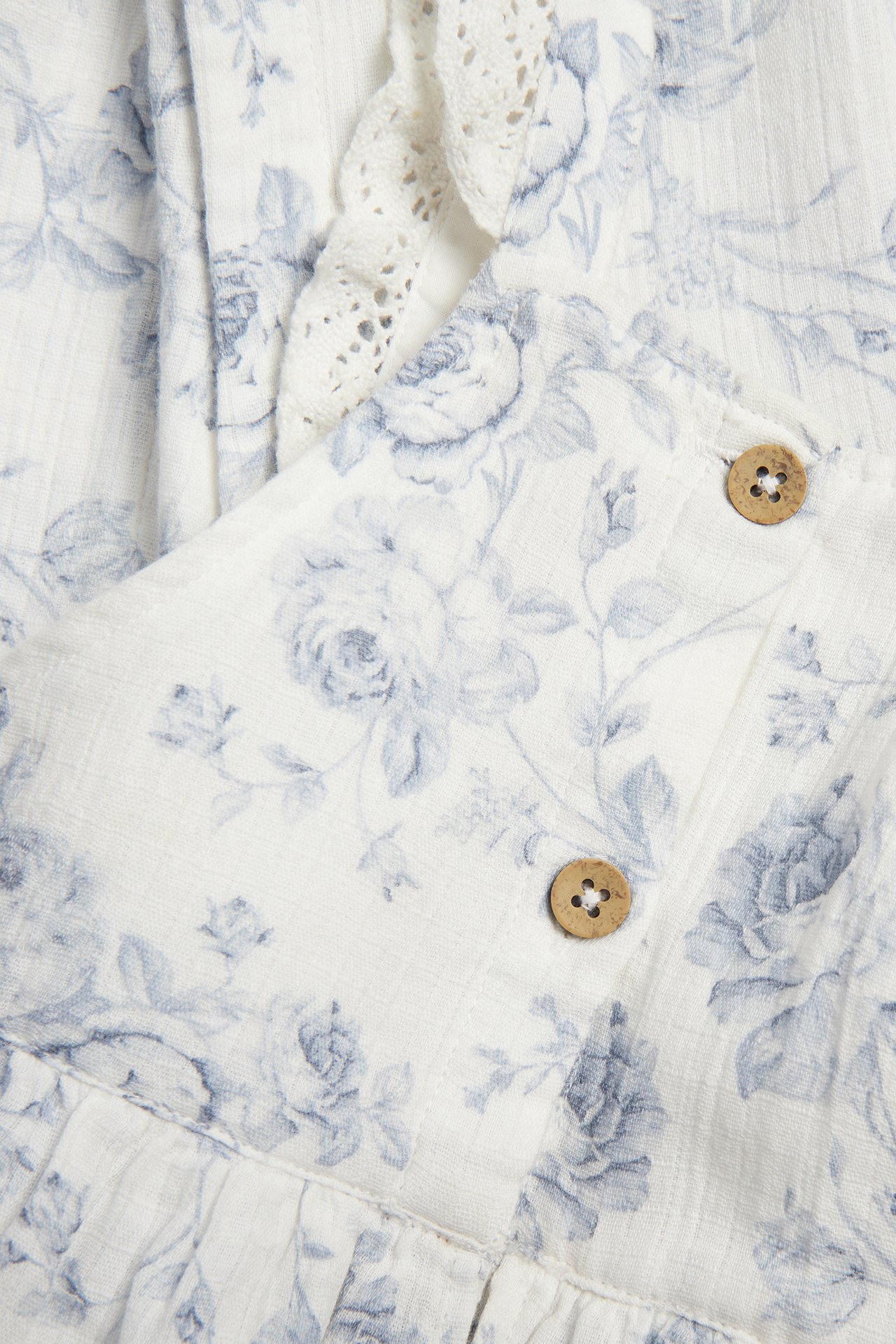 Close-up of off-white cotton dungarees with blue floral pattern, ruffles, lace trim, and wooden buttons.