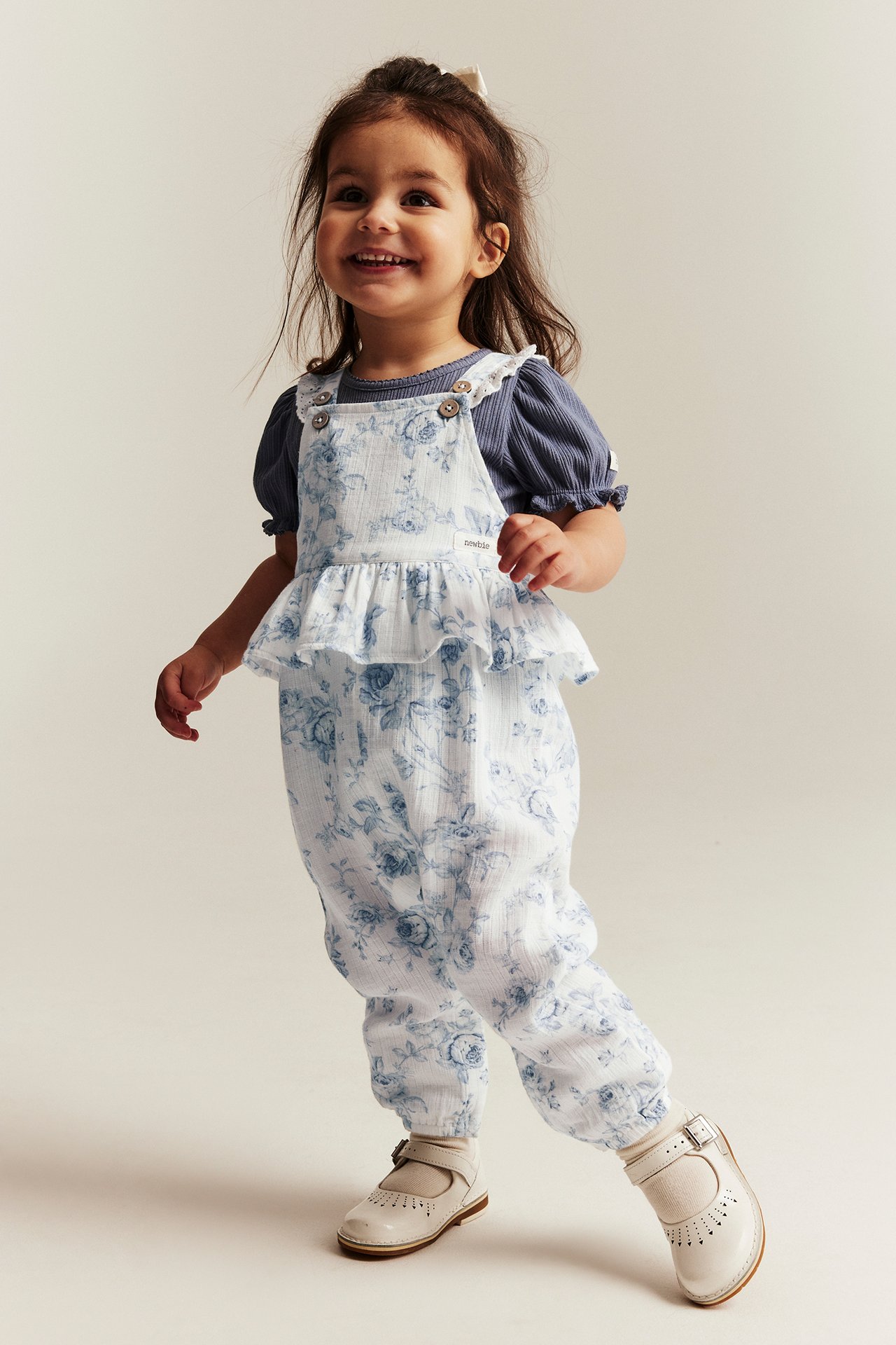 Front view: Kid in off-white floral ruffled dungarees, a blue top, and white buckle shoes.
