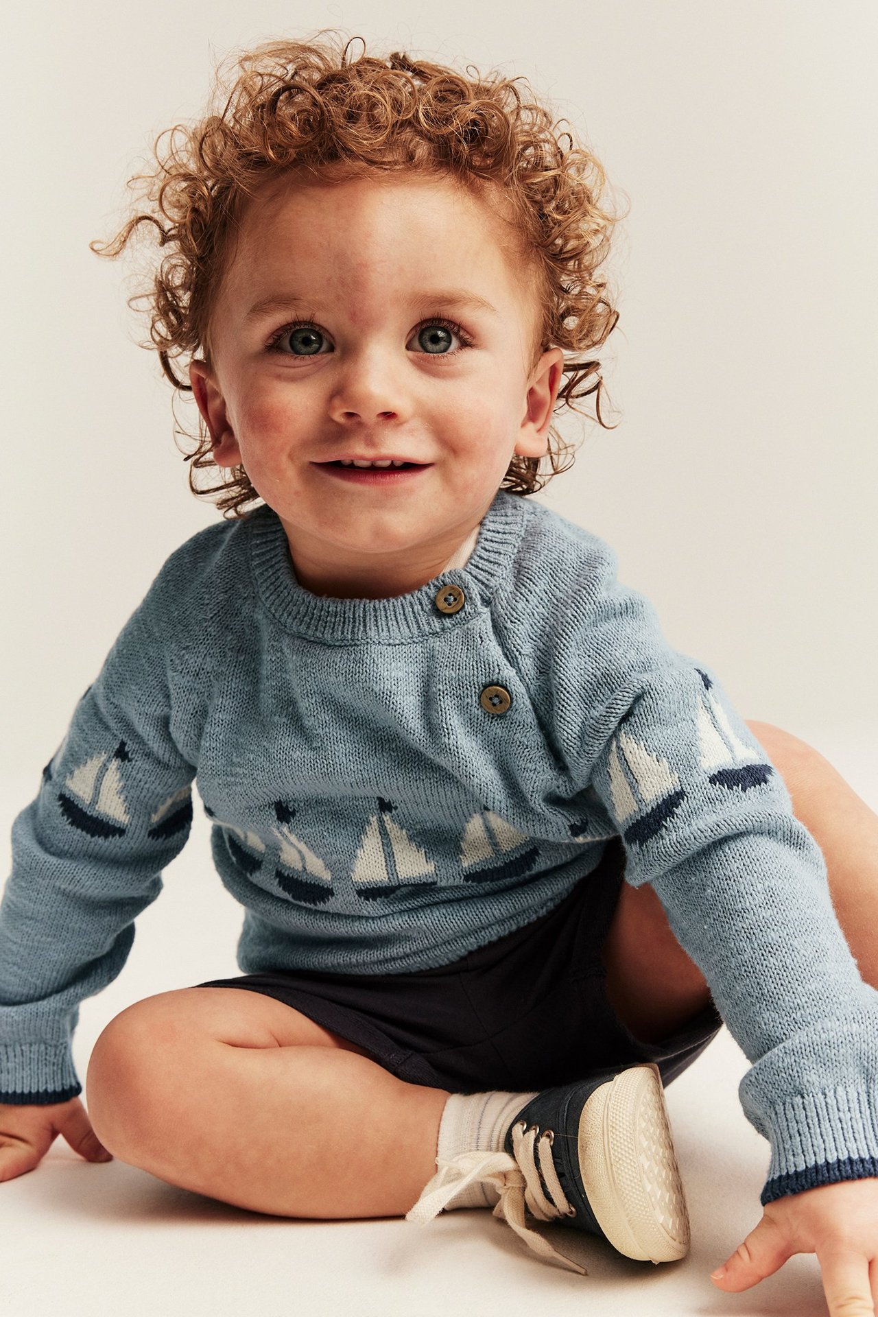 Vorderansicht eines Babys in hellblauem Baumwoll-Strickpullover mit Segelbootmuster, dunkelblauen Shorts und Schuhen.