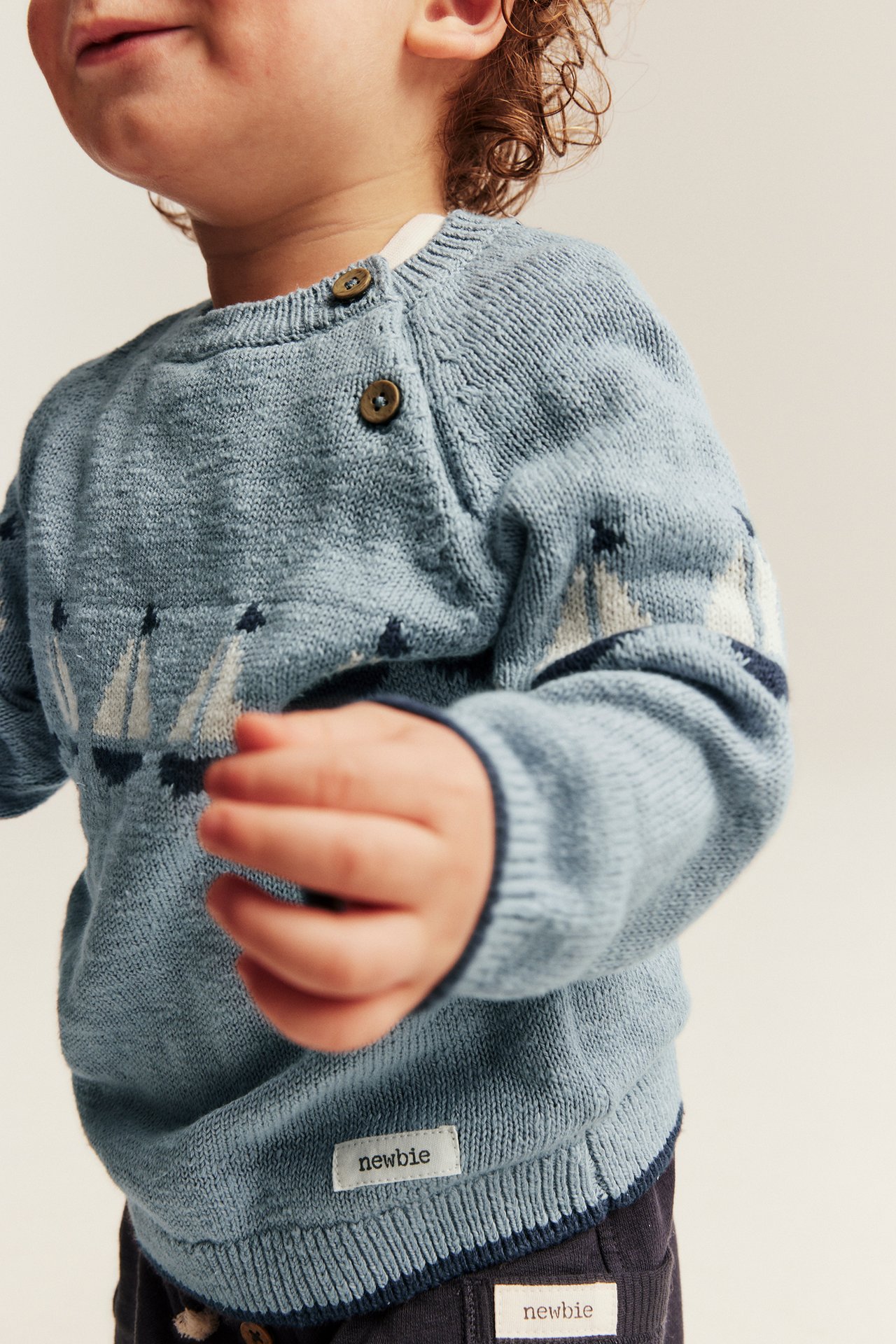 Nahaufnahme eines Babys in hellblauem gestricktem Baumwollpullover mit Segelboot-Jacquard und Schulterknöpfen.
