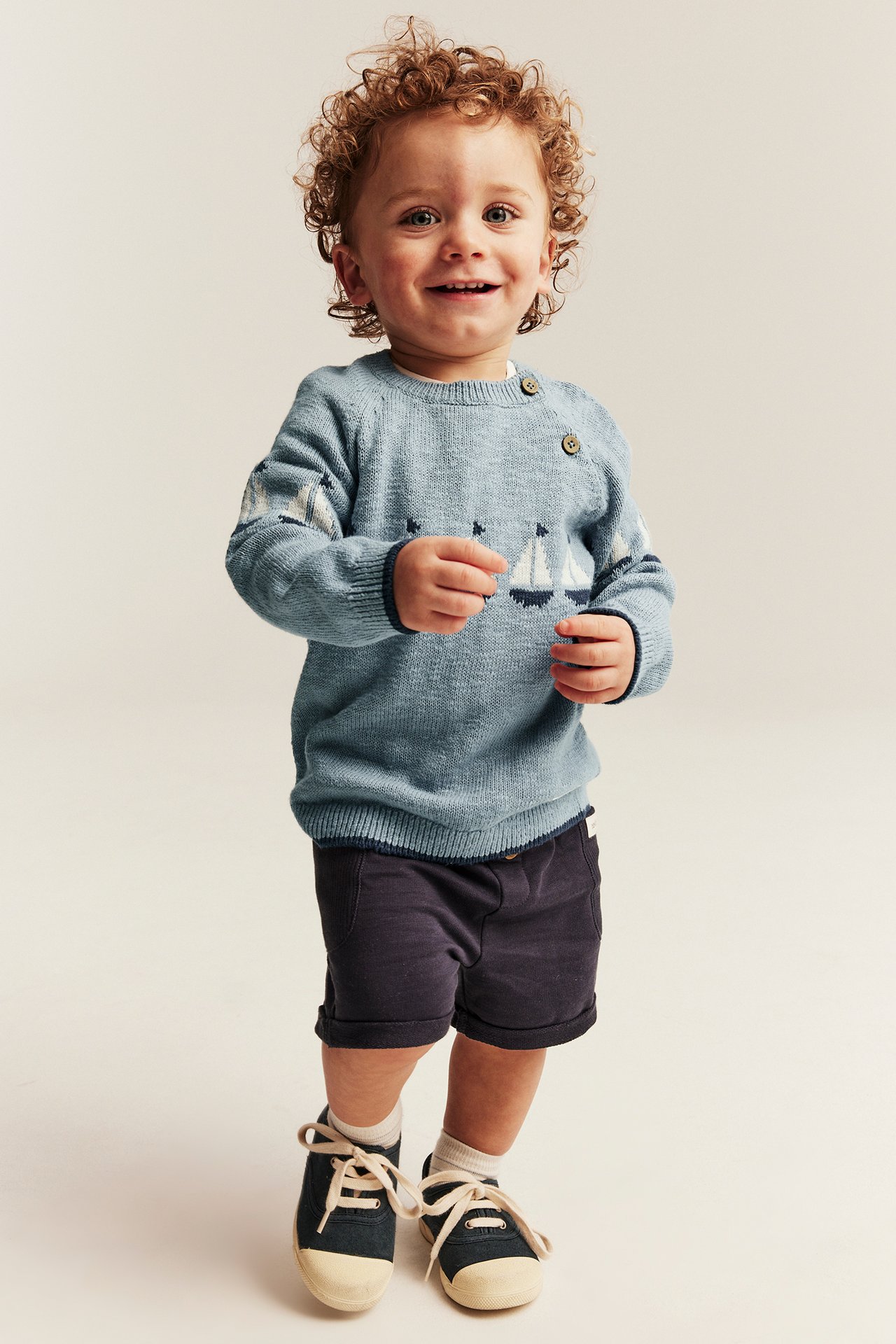 Vorderansicht: Baby trägt einen hellblauen Strickpullover mit Segelbootmuster, Schulterknöpfen und dunkelblauen Shorts.