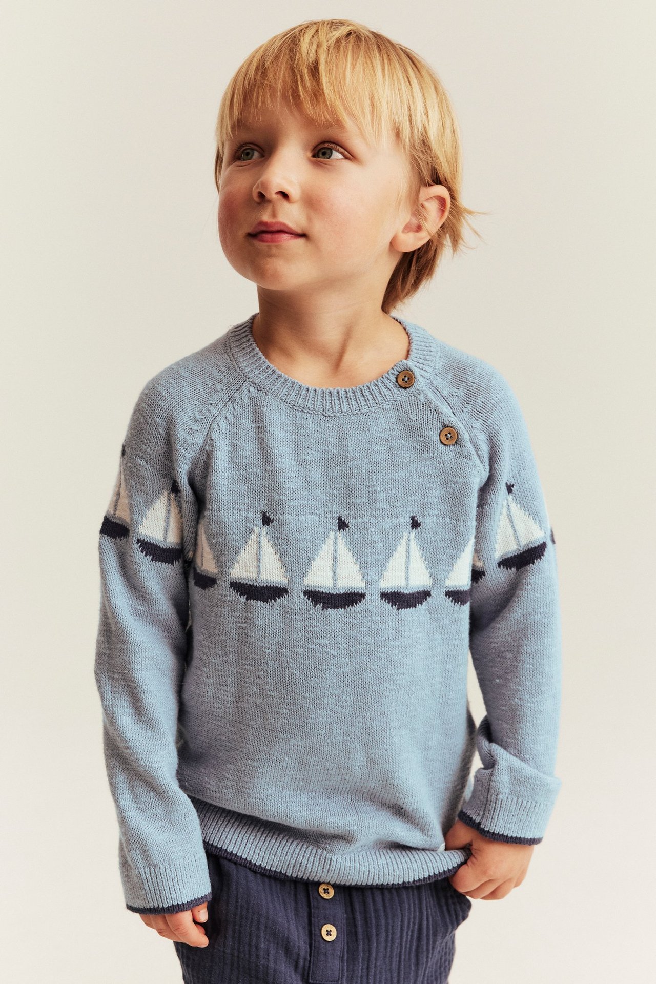 Vorderansicht: Kind trägt einen blauen Jacquard-Segelboot-Pullover mit Schulterknöpfen und dunkelblauer Hose.