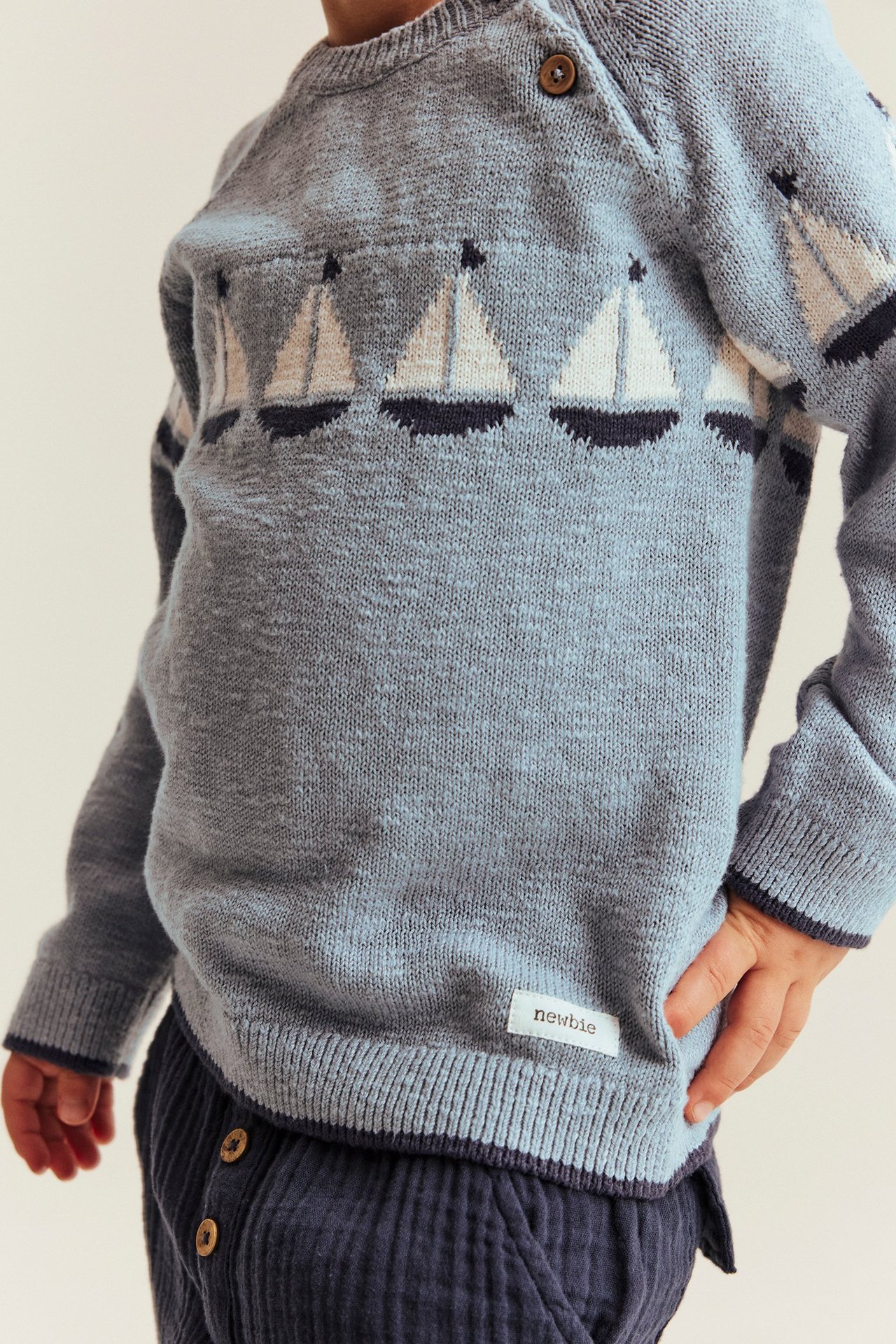 Vorderansicht eines Babys im blauen Strickpullover mit Segelboot-Jacquard, Schulterknöpfen und dunkelblauer Hose.