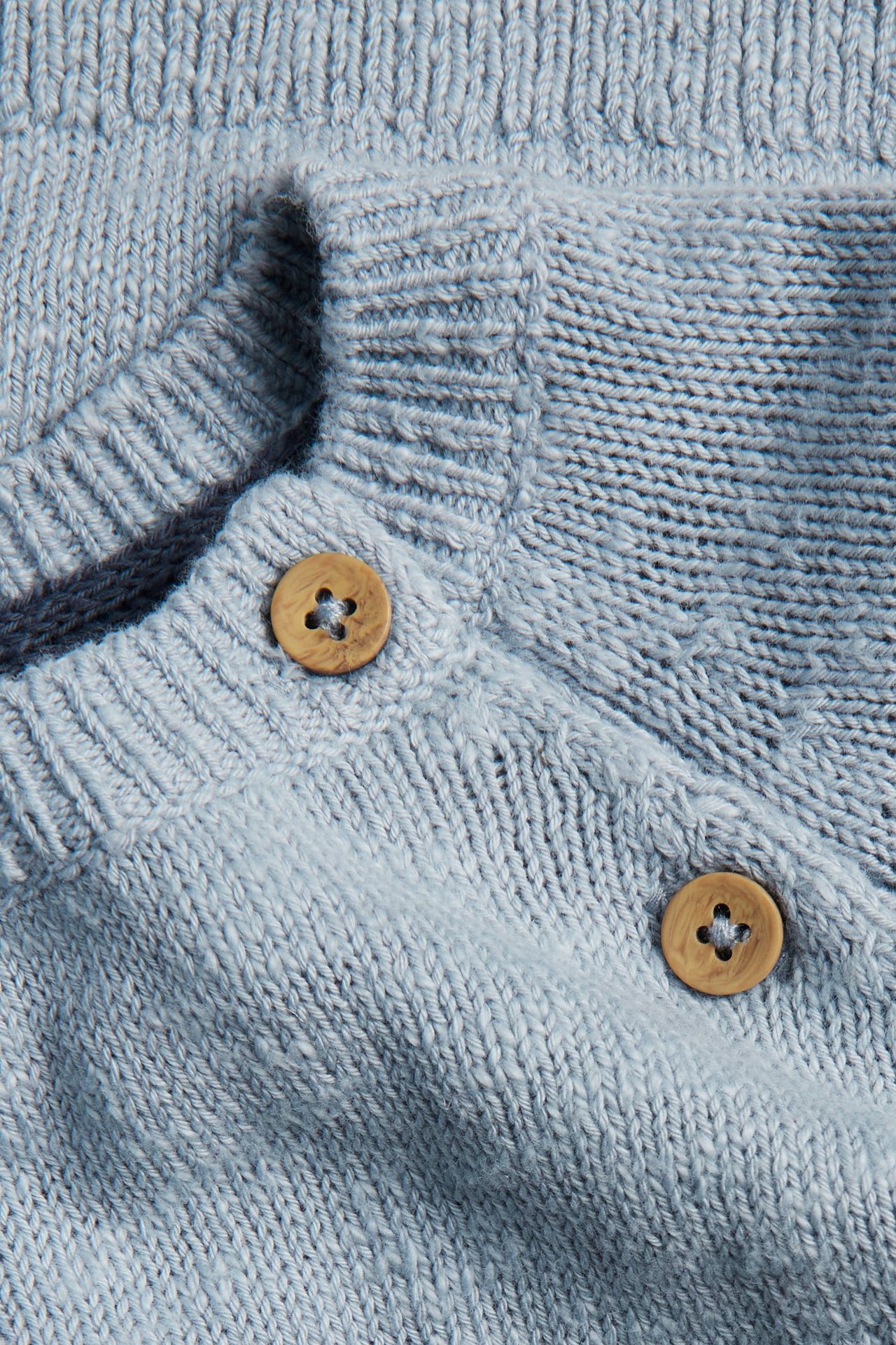 Nahaufnahme eines hellblauen Strickpullovers mit dezentem Jacquardmuster, Rippenstruktur und zwei Holzknöpfen.