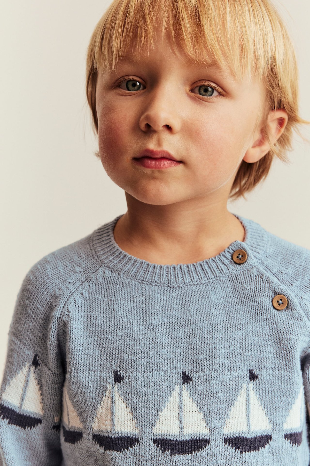Kind trägt hellblauen, gestrickten Baumwollpullover mit weißem Segelboot-Jacquardmuster und zwei Schulterknöpfen.