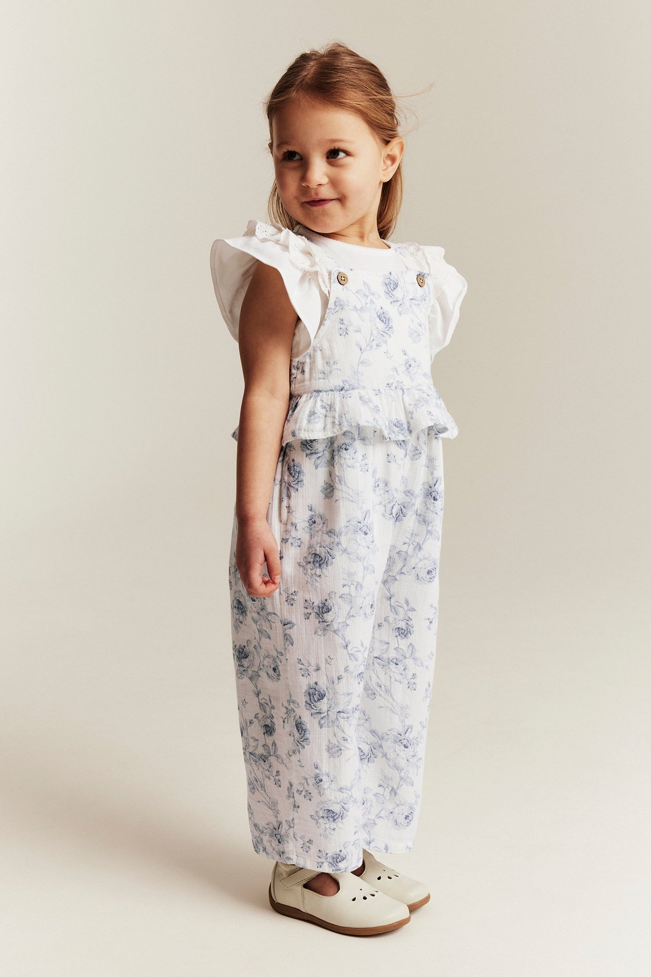 Kind trägt off-weißen floralen Jumpsuit mit Rüschensaum, verstellbaren Trägern, weißem T-Shirt und weißen Schuhen.