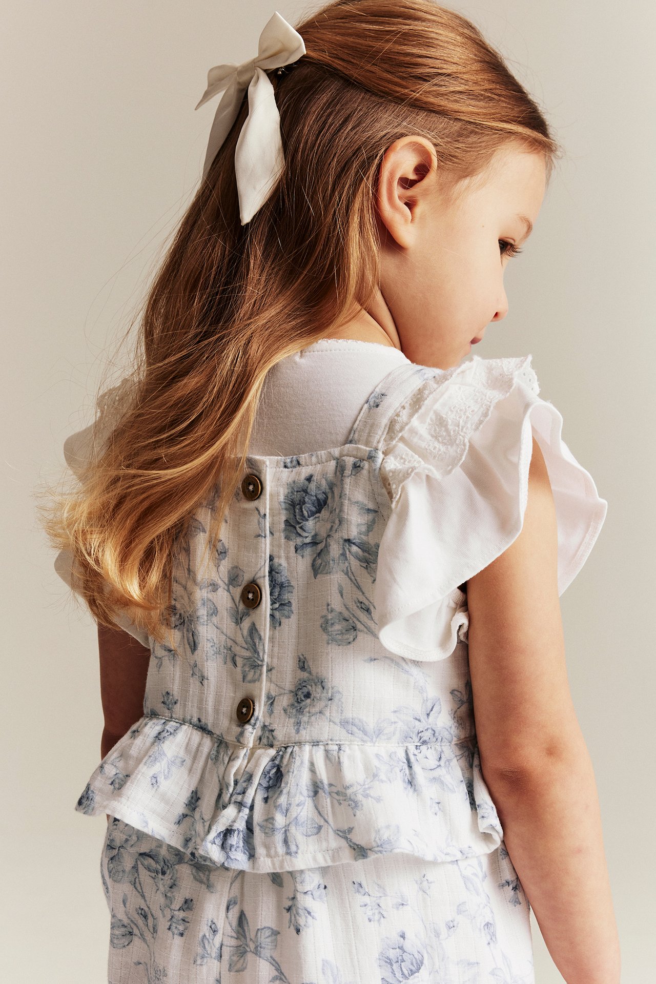 Rückenansicht: Kind in off-weißem Blumen-Overall mit Rüschen, Knöpfen, über weißem T-Shirt, weißer Schleife.