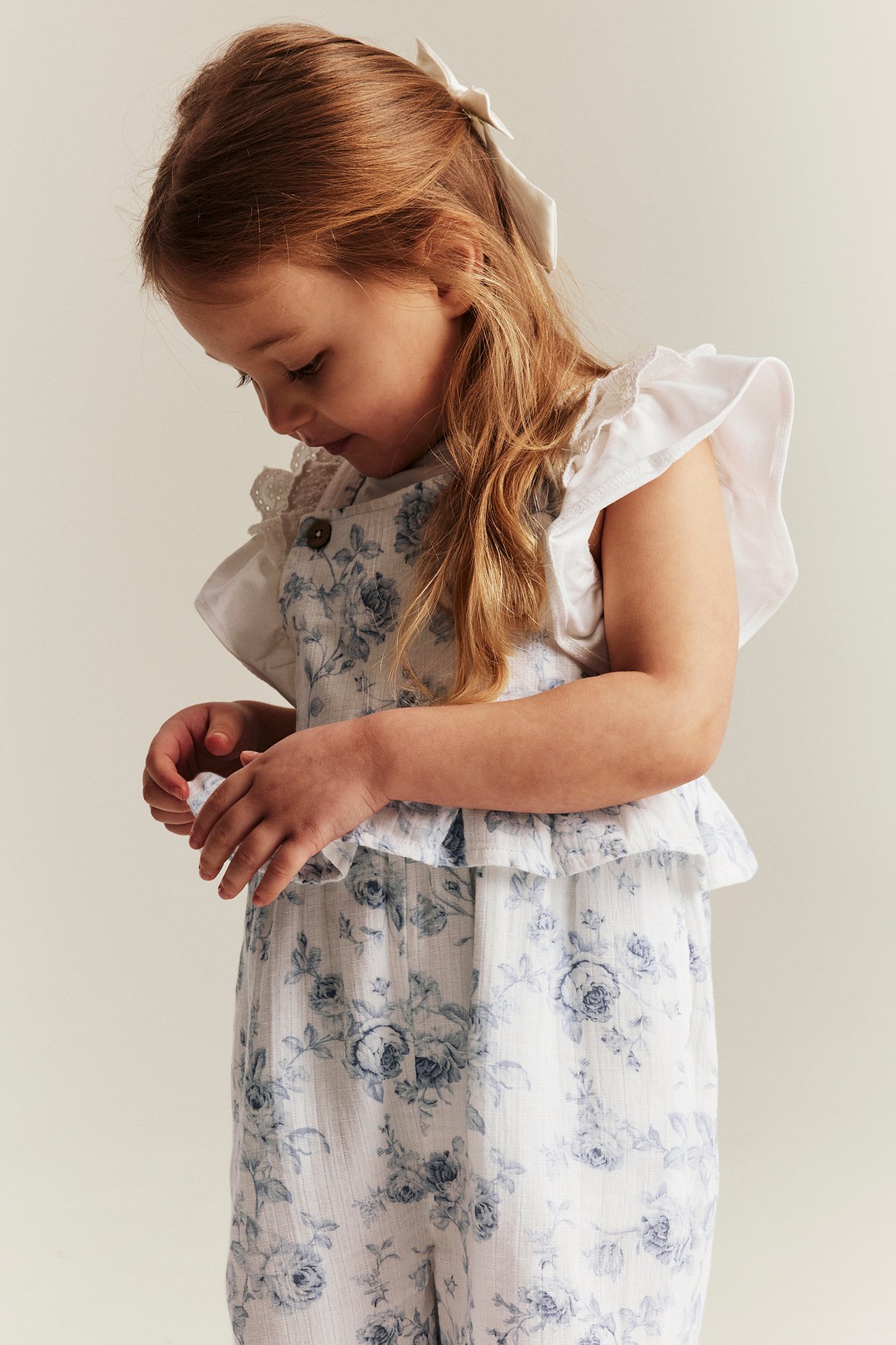 Vorderansicht: Kind trägt einen cremeweißen floralen Overall mit blauem Muster, Taillenrüsche und weißem Oberteil.