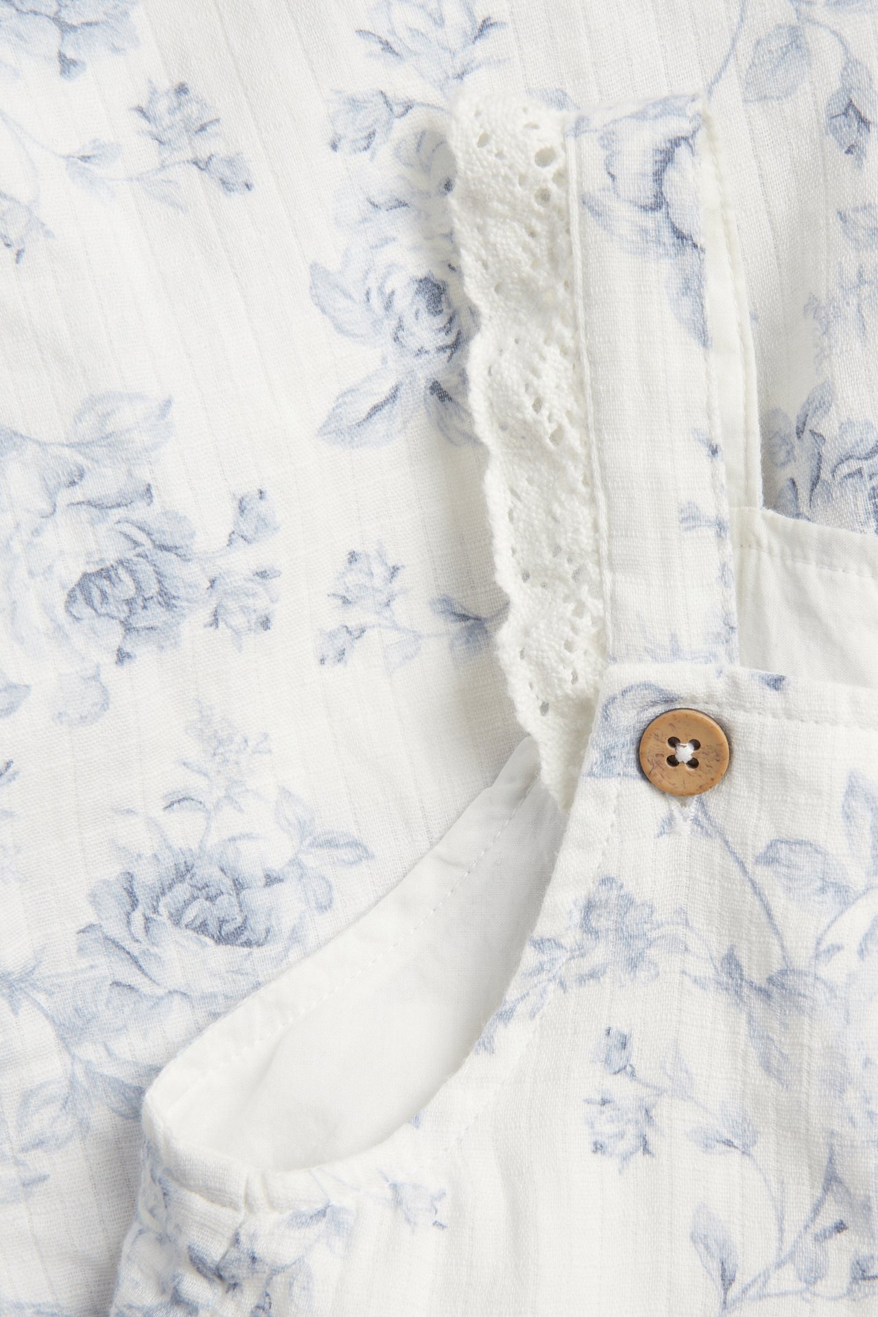 Nahaufnahme eines off-white Baumwoll-Blumen-Jumpsuits mit hellblauem Muster, Spitzenrüsche und Holzknopf.