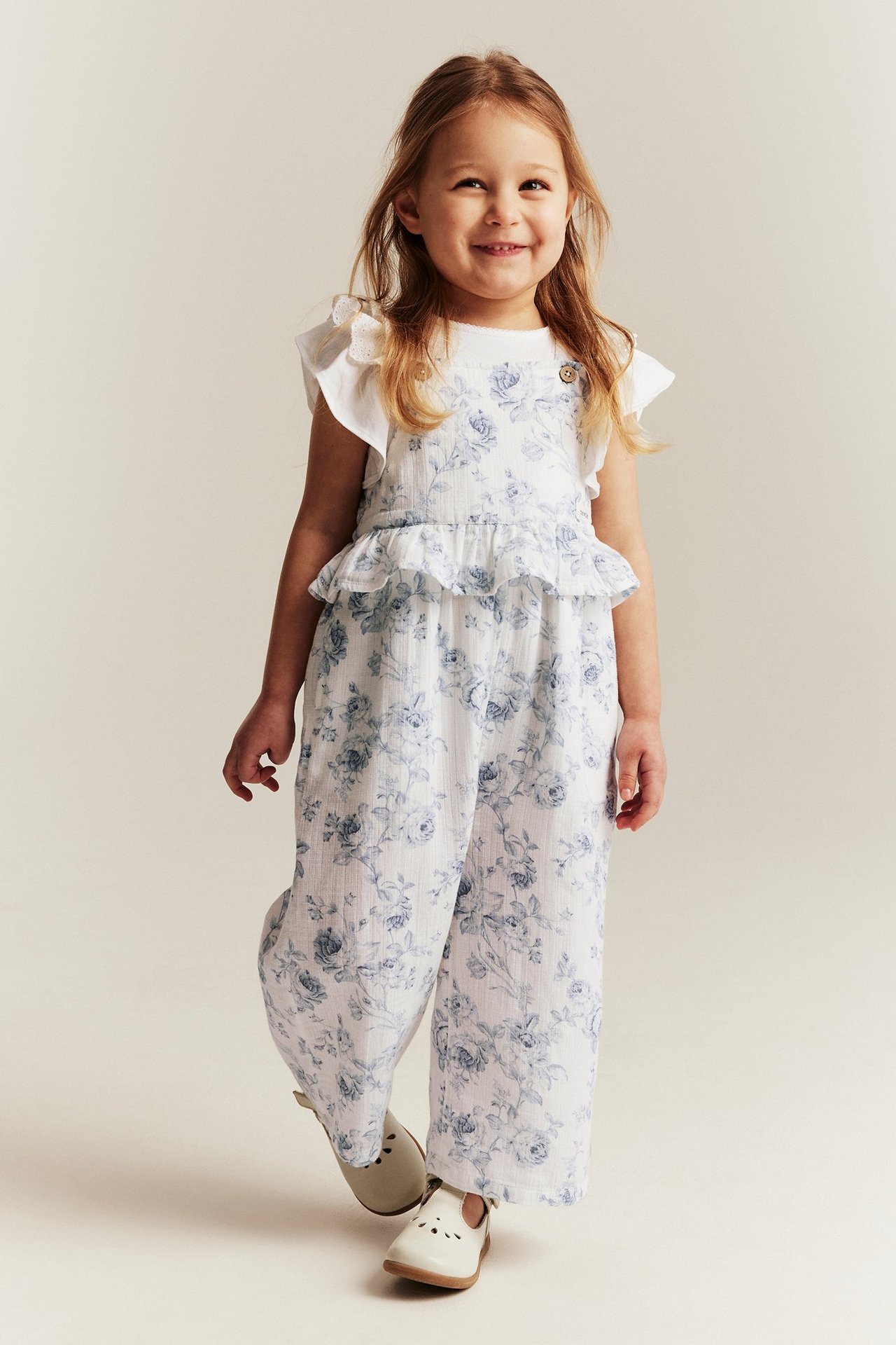 Front view: A smiling kid wears an off-white floral cotton jumpsuit with ruffled straps and light shoes.