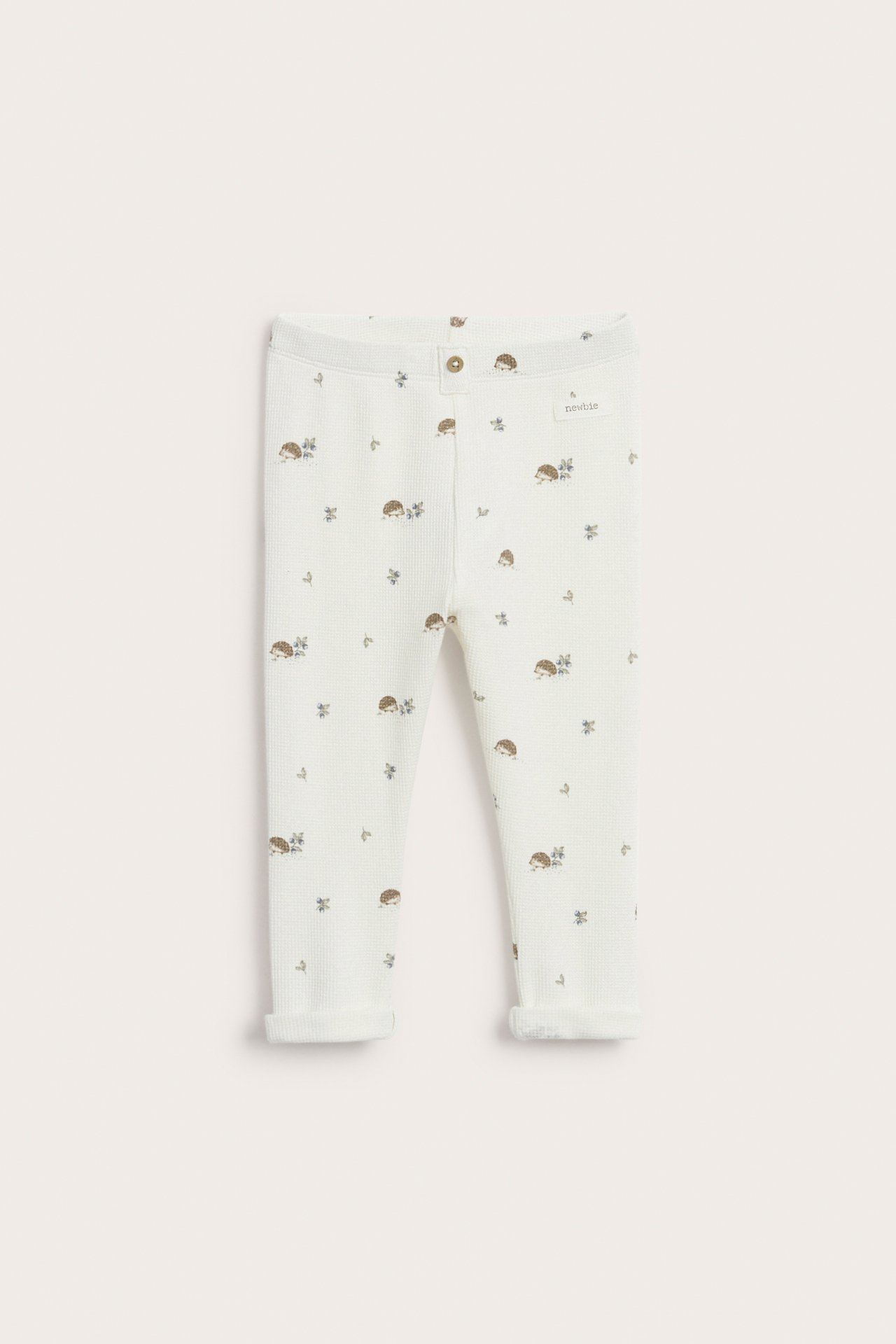Front view of off-white waffle-knit baby leggings with hedgehog pattern, decorative front button, and rolled-up cuffs.