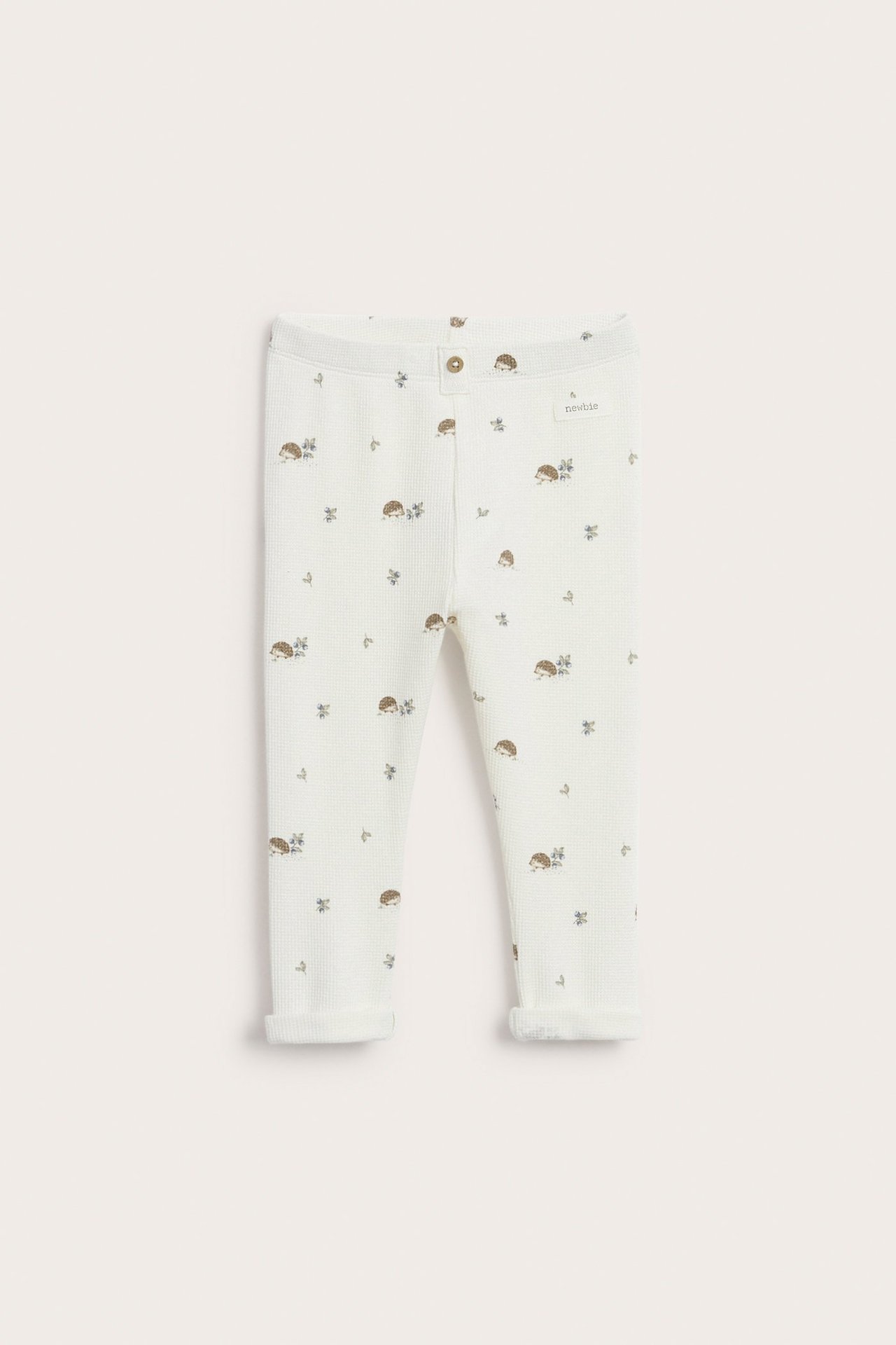 Vorderansicht von cremeweißen Waffelstrick-Baby-Leggings mit Igelmuster, Zierknopf vorne und umgekrempelten Beinabschlüssen.