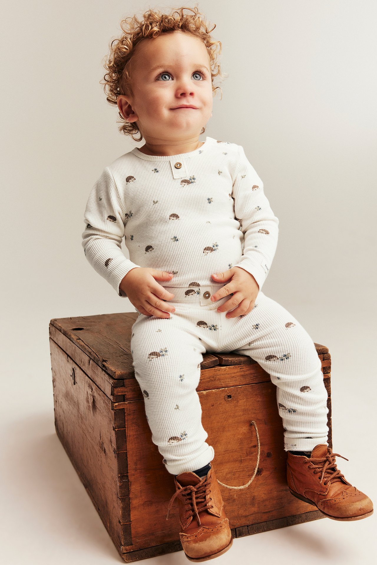 Front view of a baby in an off-white animal-patterned waffle-knit long-sleeved bodysuit, matching trousers, and brown shoes.