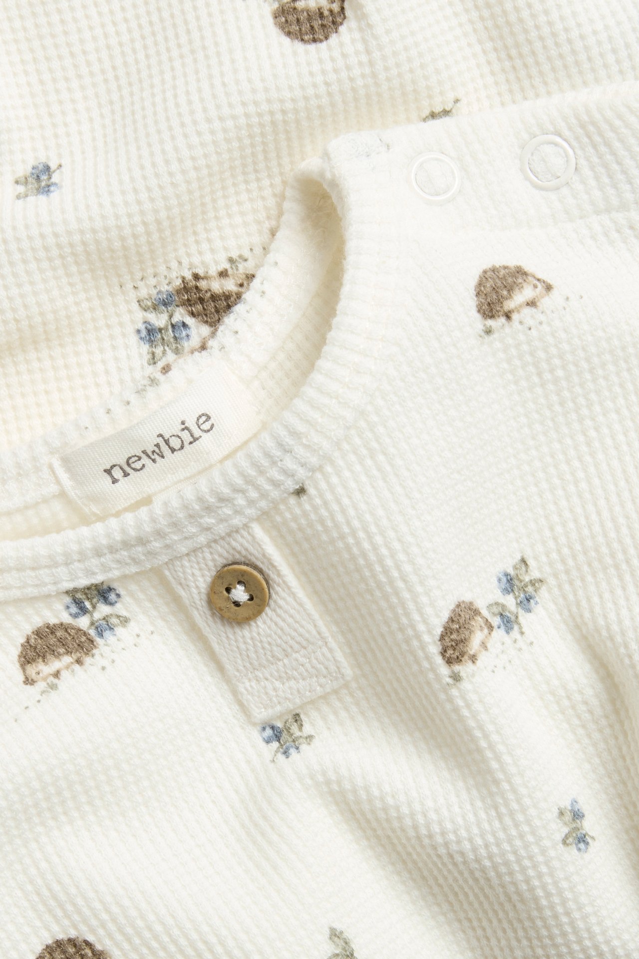 Close-up: off-white waffle knit baby bodysuit with hedgehog and floral print, decorative button, and shoulder snaps.