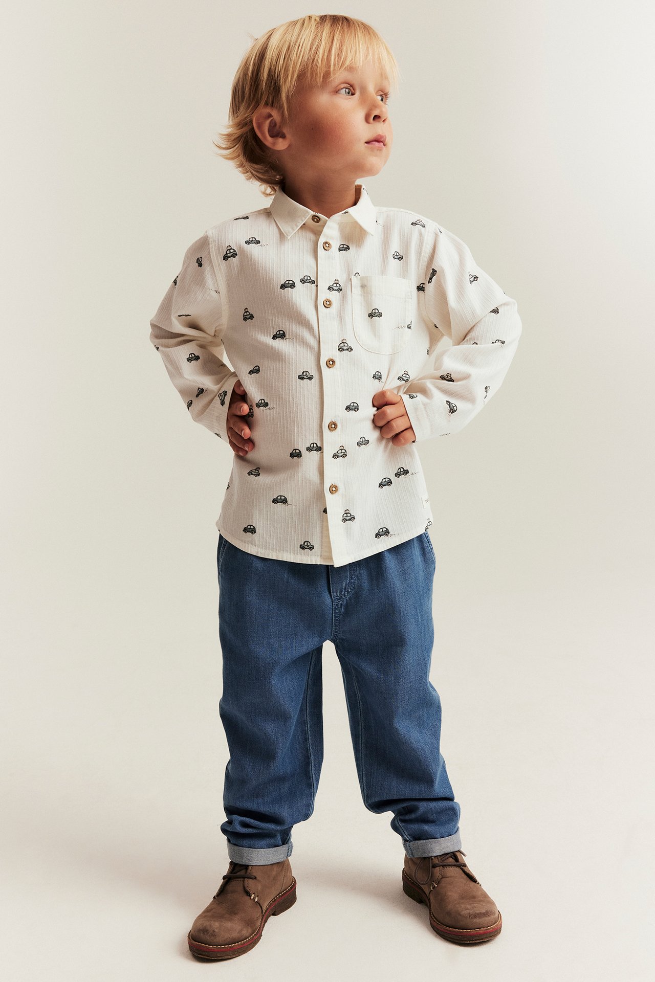 Front view of a kid wearing an off-white cotton car-patterned shirt, blue jeans, and brown lace-up boots.