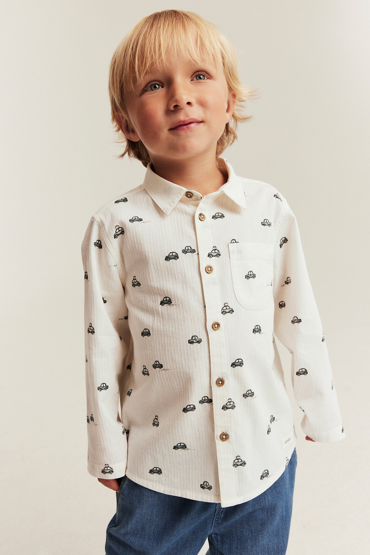 Front view of a kid wearing an offwhite cotton shirt with a black car pattern and blue jeans.