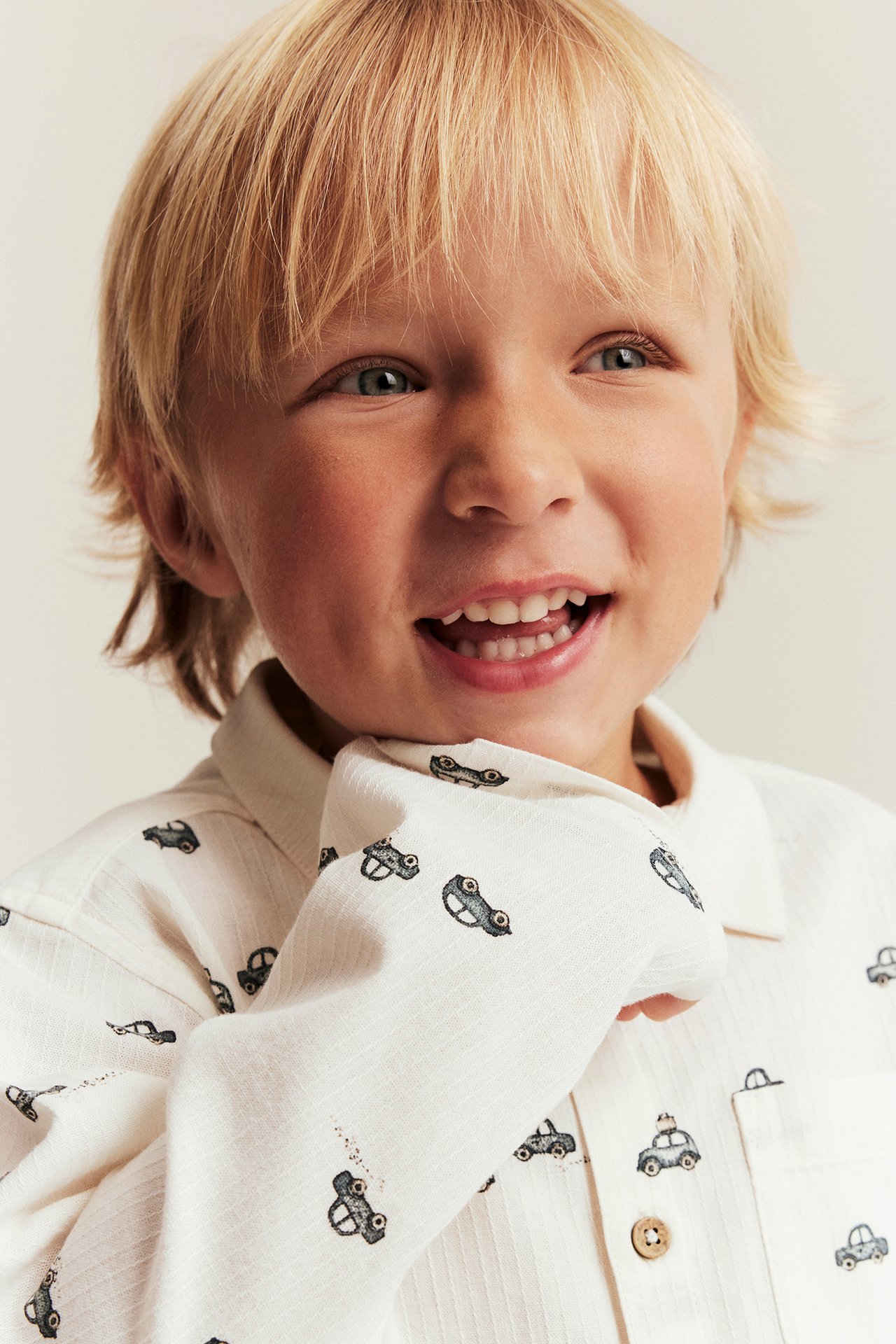 Close-up of a smiling kid wearing an off-white woven shirt with a car pattern, collar, and front buttons.
