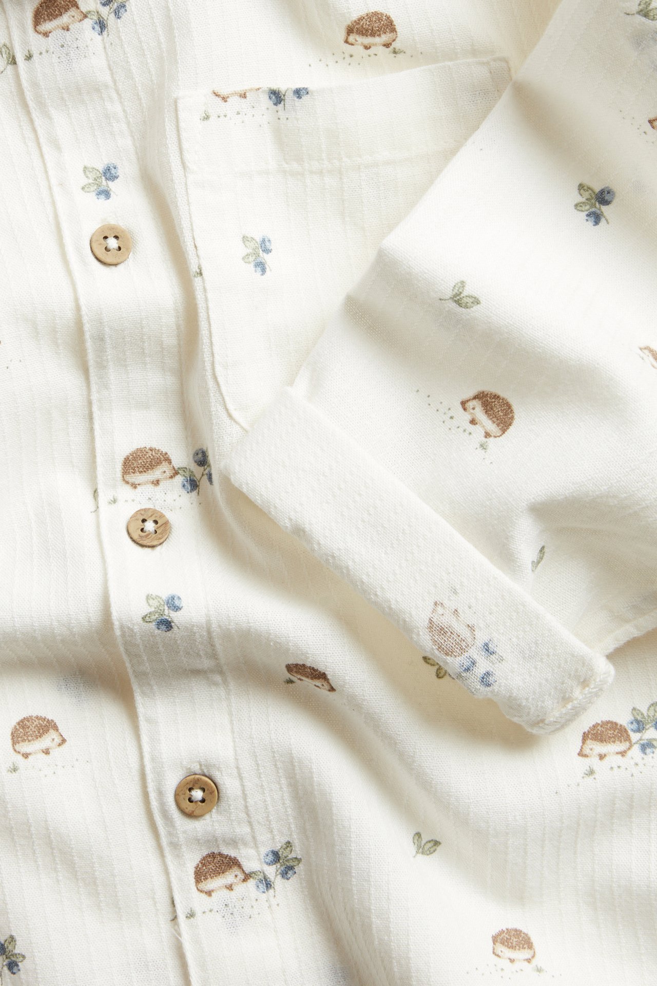 Close-up of an off-white cotton kid's shirt with hedgehog and blueberry pattern, wooden buttons, and rolled-up sleeve.