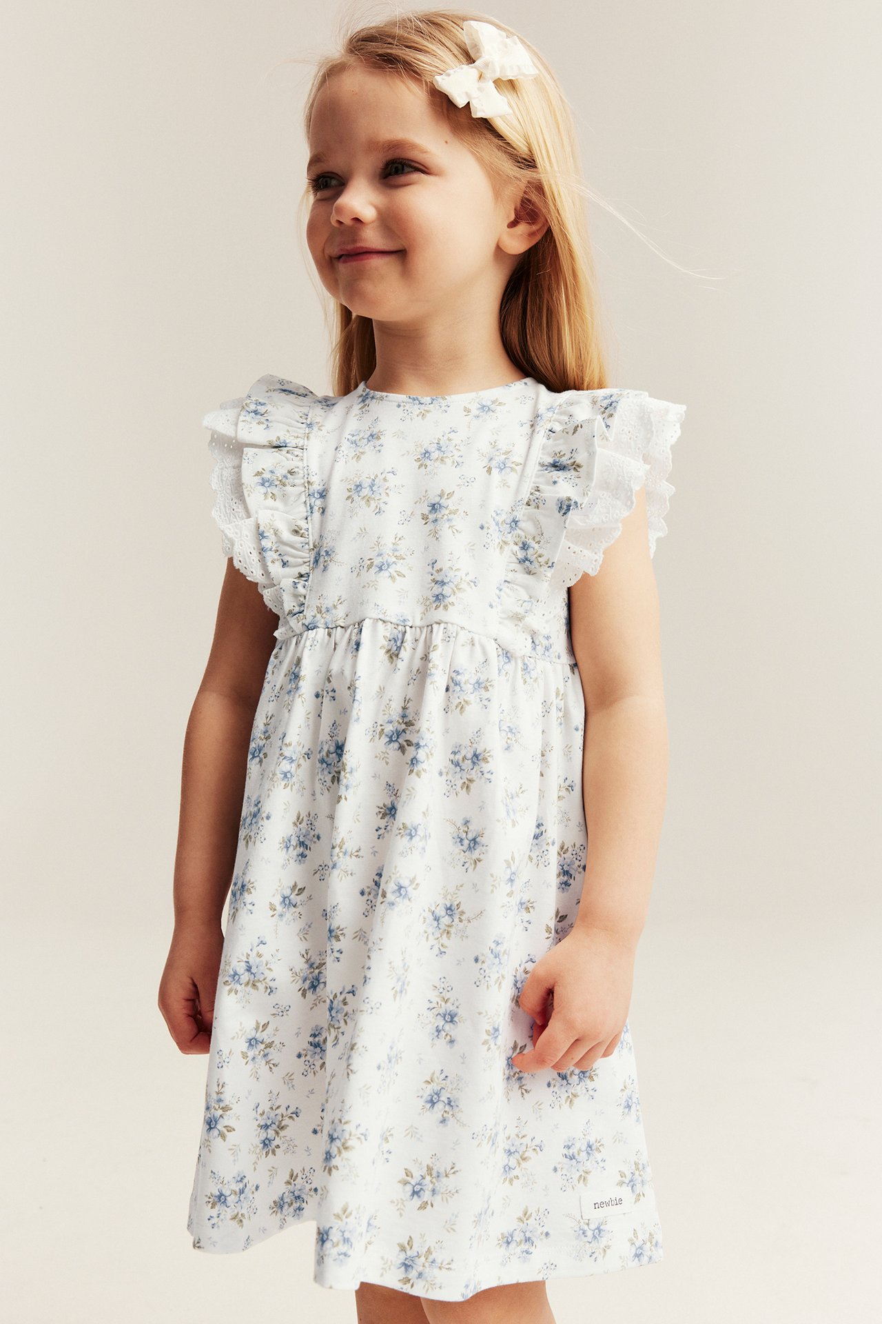 Front view of a kid in an offwhite floral jersey dress with ruffled, lace-trimmed shoulders and a white bow hair clip.
