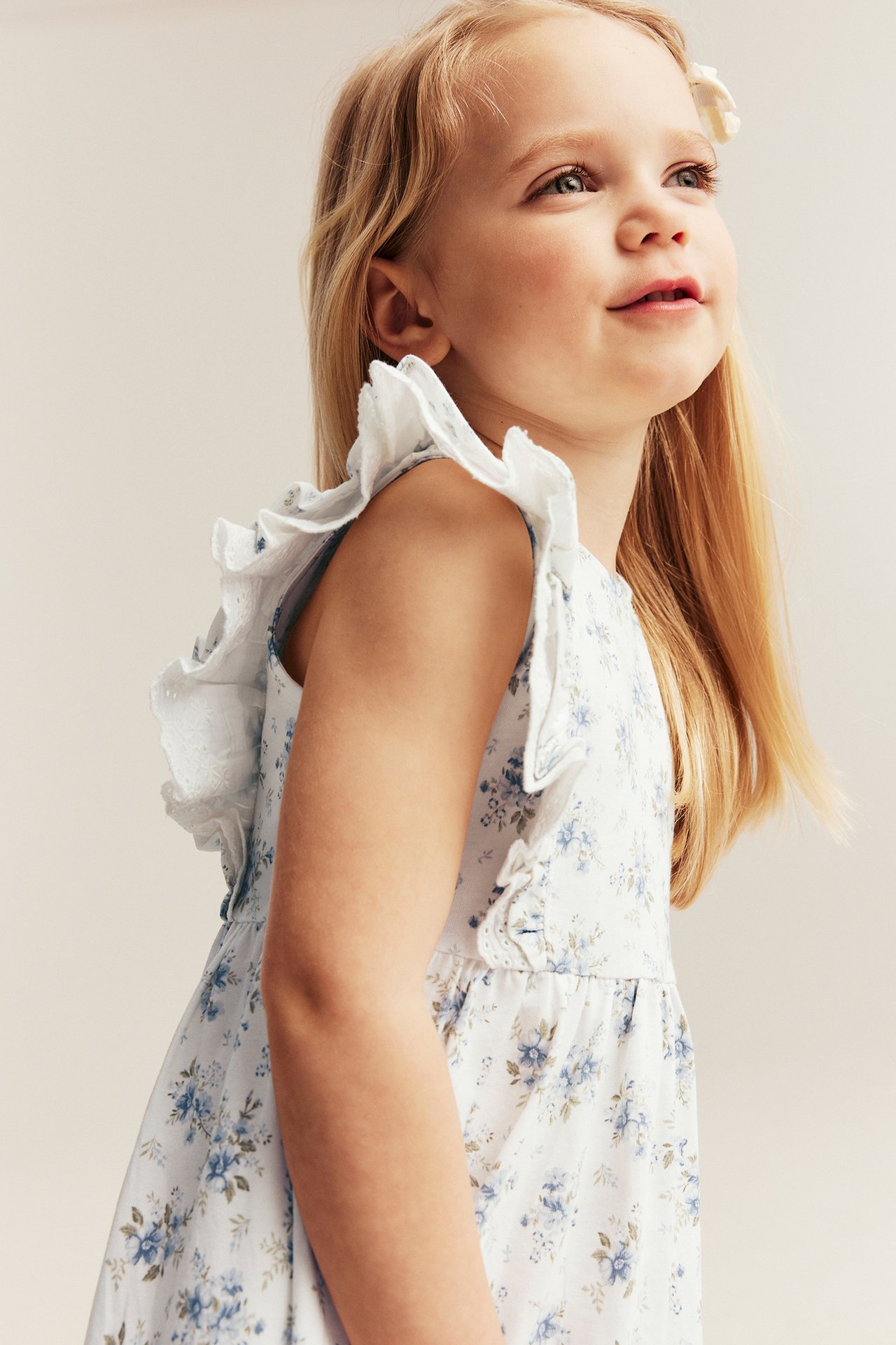Seitenansicht eines Kindes in einem cremeweißen geblümten Baumwoll-Jerseykleid mit Rüschen-Spitzenschultern.