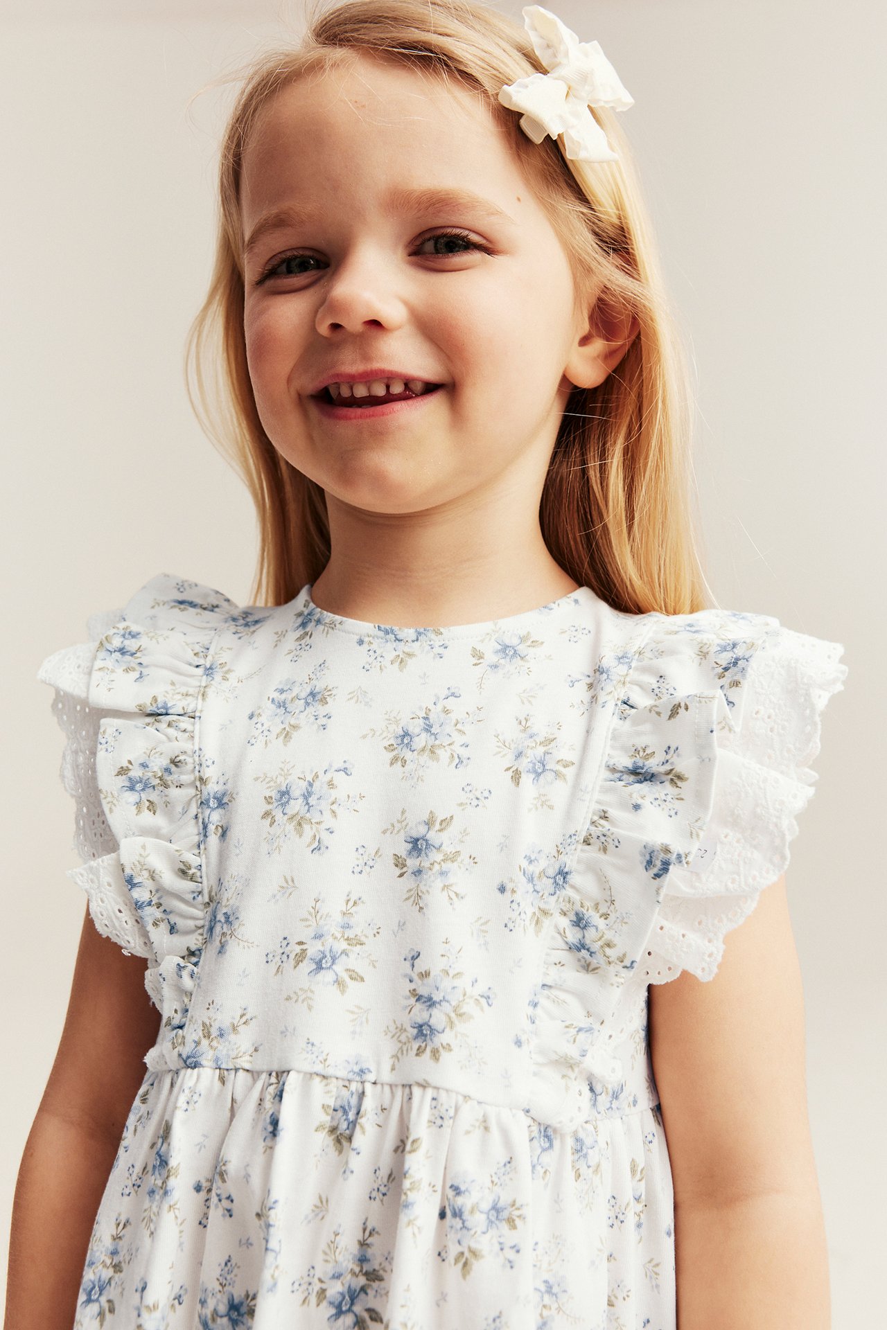 Vorderansicht: Kind trägt ein cremefarbenes Blumen-Jerseykleid mit Rüschen-Spitzenschultern und weißer Haarschleife.