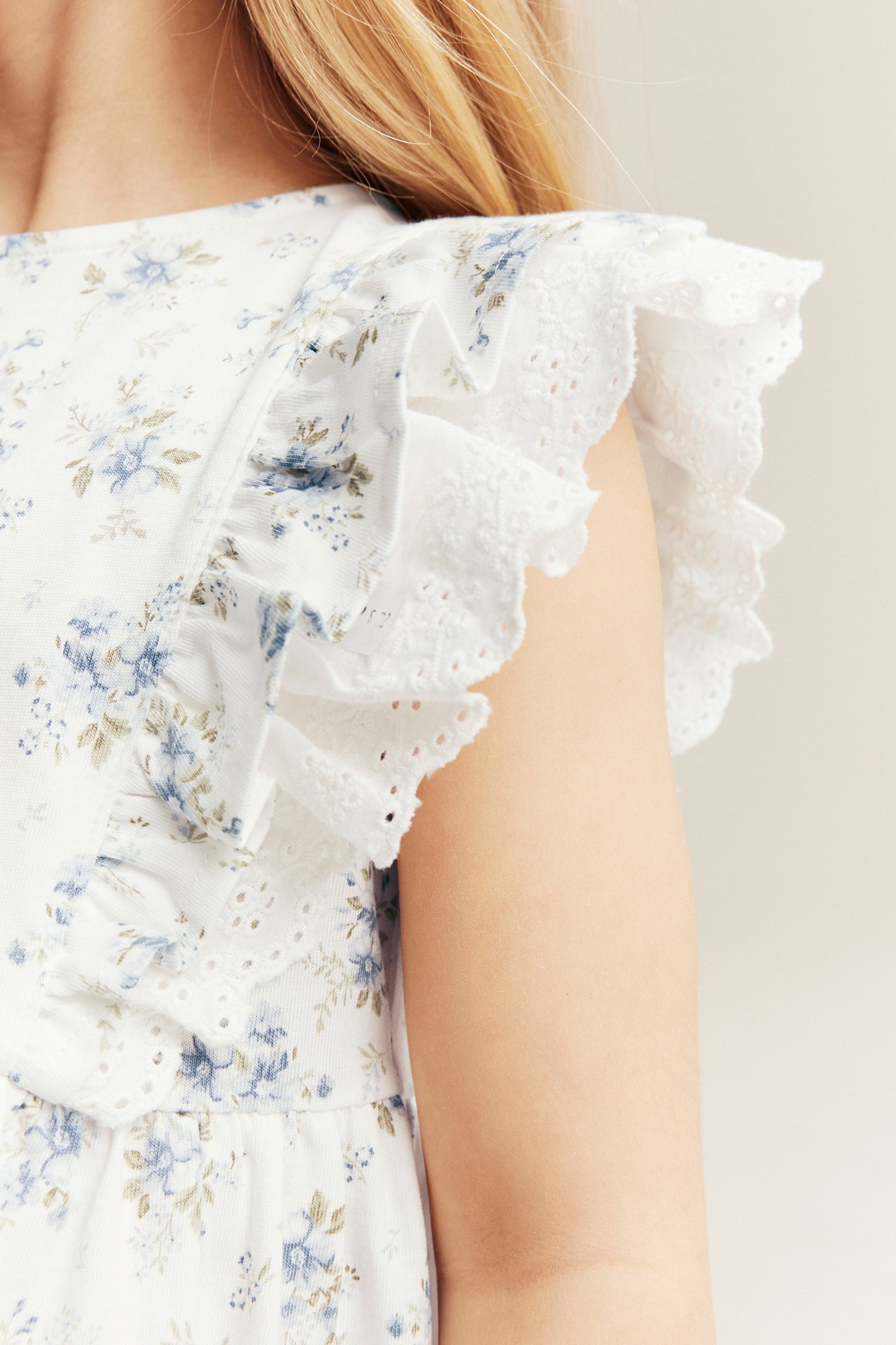 Nahaufnahme: Kind trägt cremeweißes geblümtes Jerseykleid mit Rüschen und Spitzendetails.
