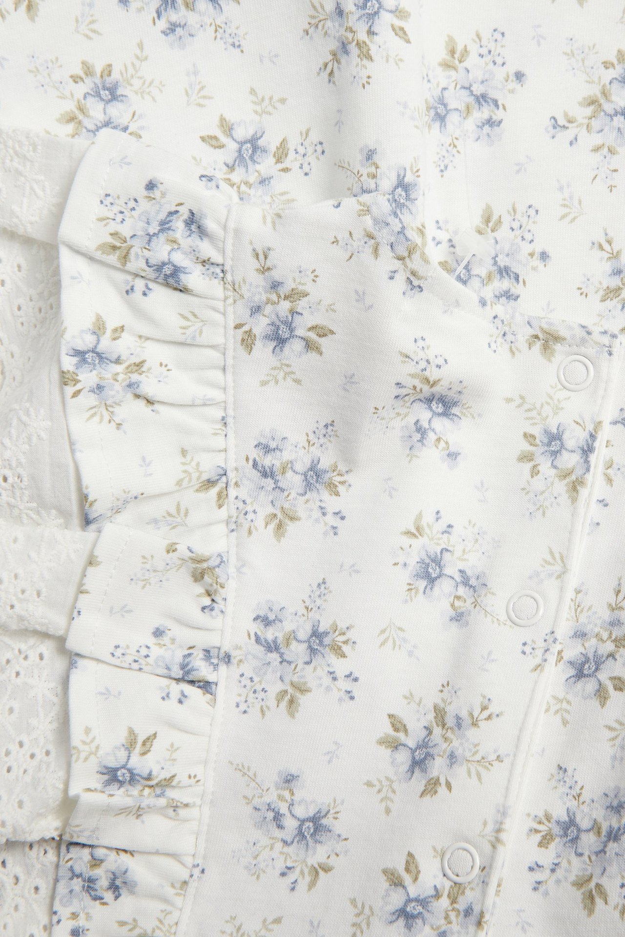 Nahaufnahme eines cremeweißen Baby-Jerseykleides mit blauem Blumenmuster, Rüschen, weißer Spitze und Druckknöpfen.