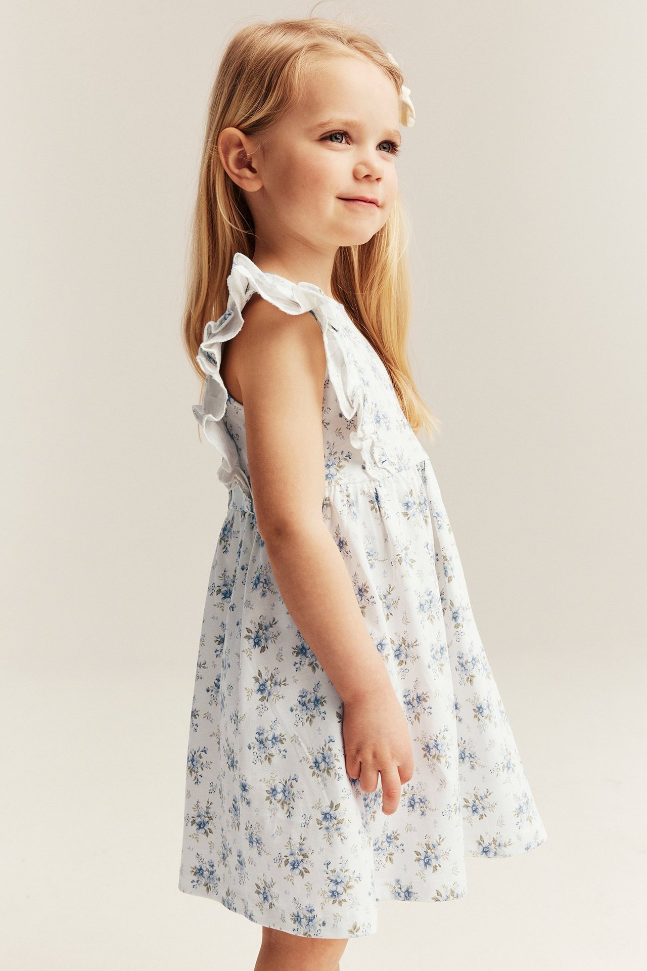 Side view of a kid in an offwhite cotton dress with blue floral print, ruffled lace shoulders.