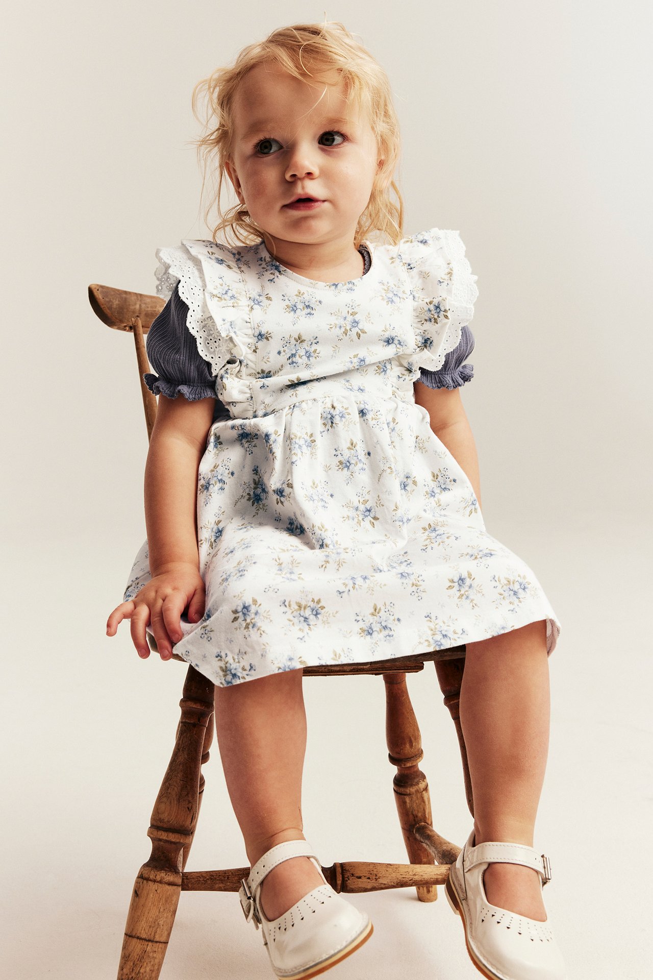 Baby wears an offwhite floral dress with lace ruffles, a blue top, and white Mary Jane shoes.