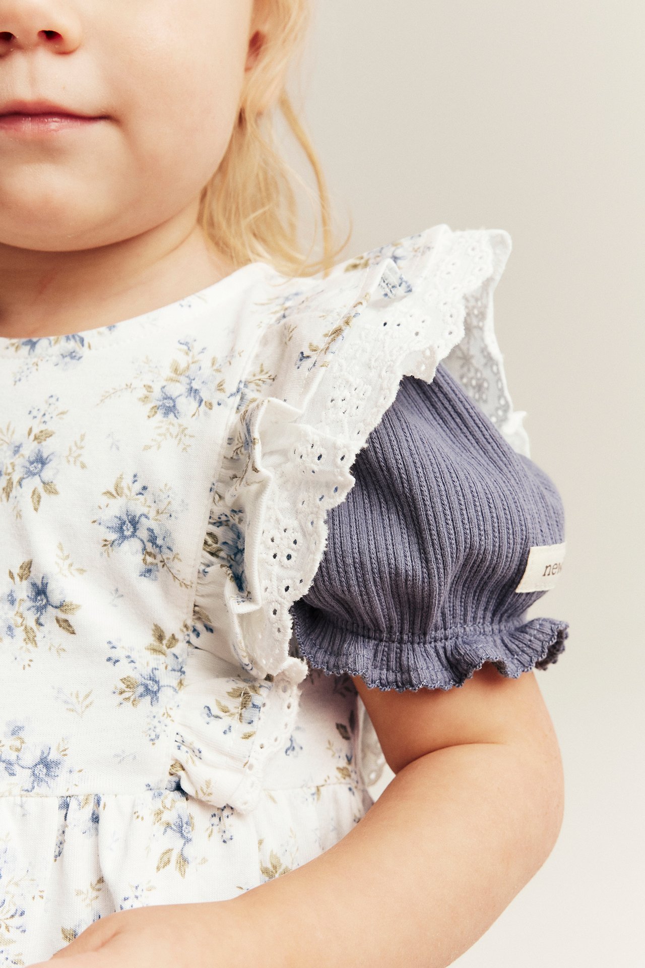 Nahaufnahme eines Kindes in einem cremefarbenen geblümten Jerseykleid mit Spitzenrüschen über einem gerippten lila-grauen Oberteil.