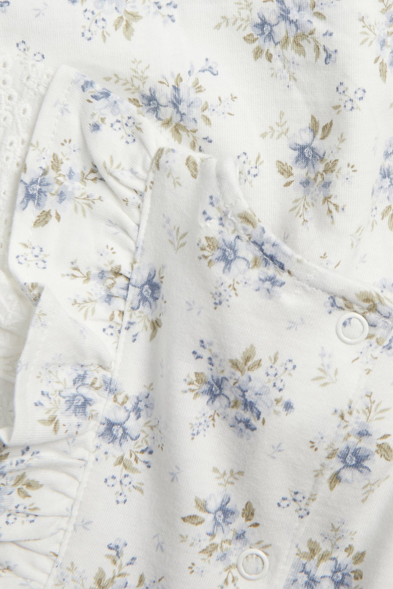 Close-up of an off-white cotton jersey floral dress with blue flowers, ruffles, lace details, and white snap buttons.