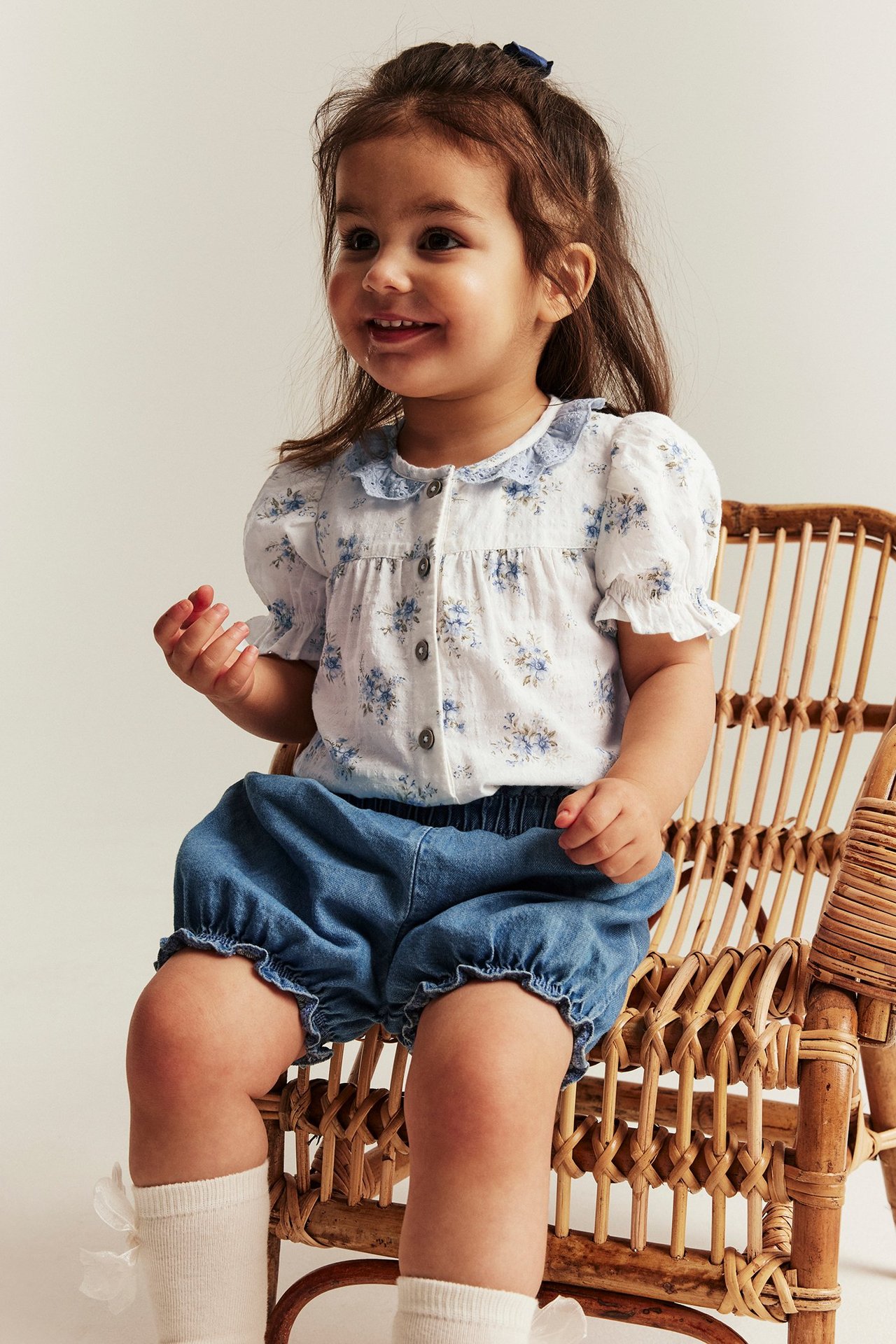 Vorderansicht eines Babymädchens in einem cremeweißen geblümten Kurzarmbody, blauen Shorts und weißen Socken.