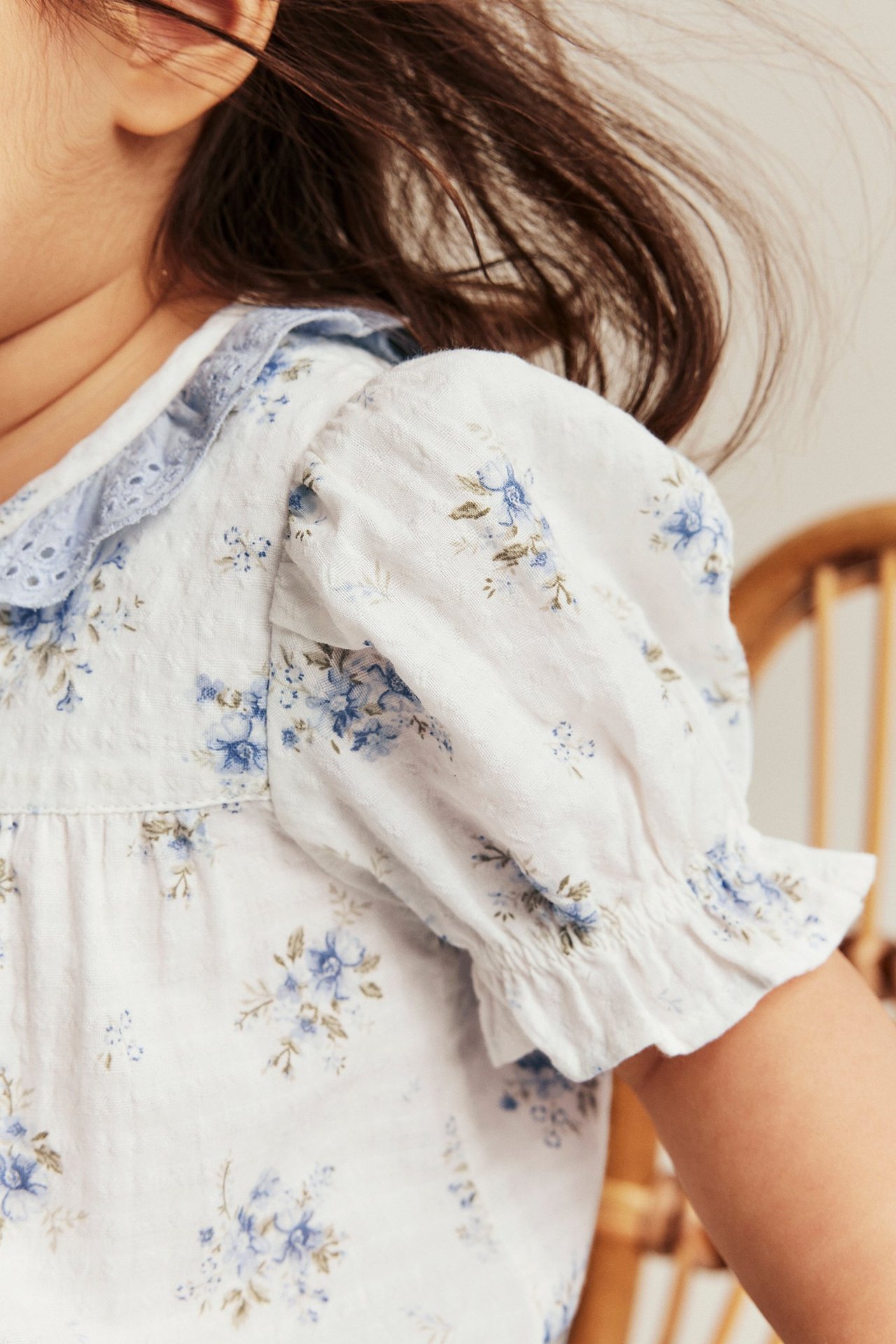 Nahaufnahme: Kind trägt einen cremefarbenen Blumen-Baumwollbody mit blauem Spitzenrüschen und Puffärmeln.