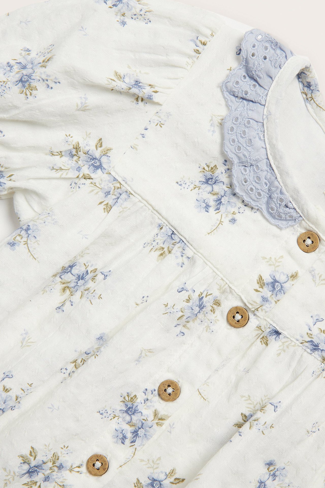 Nahaufnahme cremeweißen Baby-Bodys mit blauem Blumenmuster, hellblauem Spitzenkragen und Holzknöpfen.