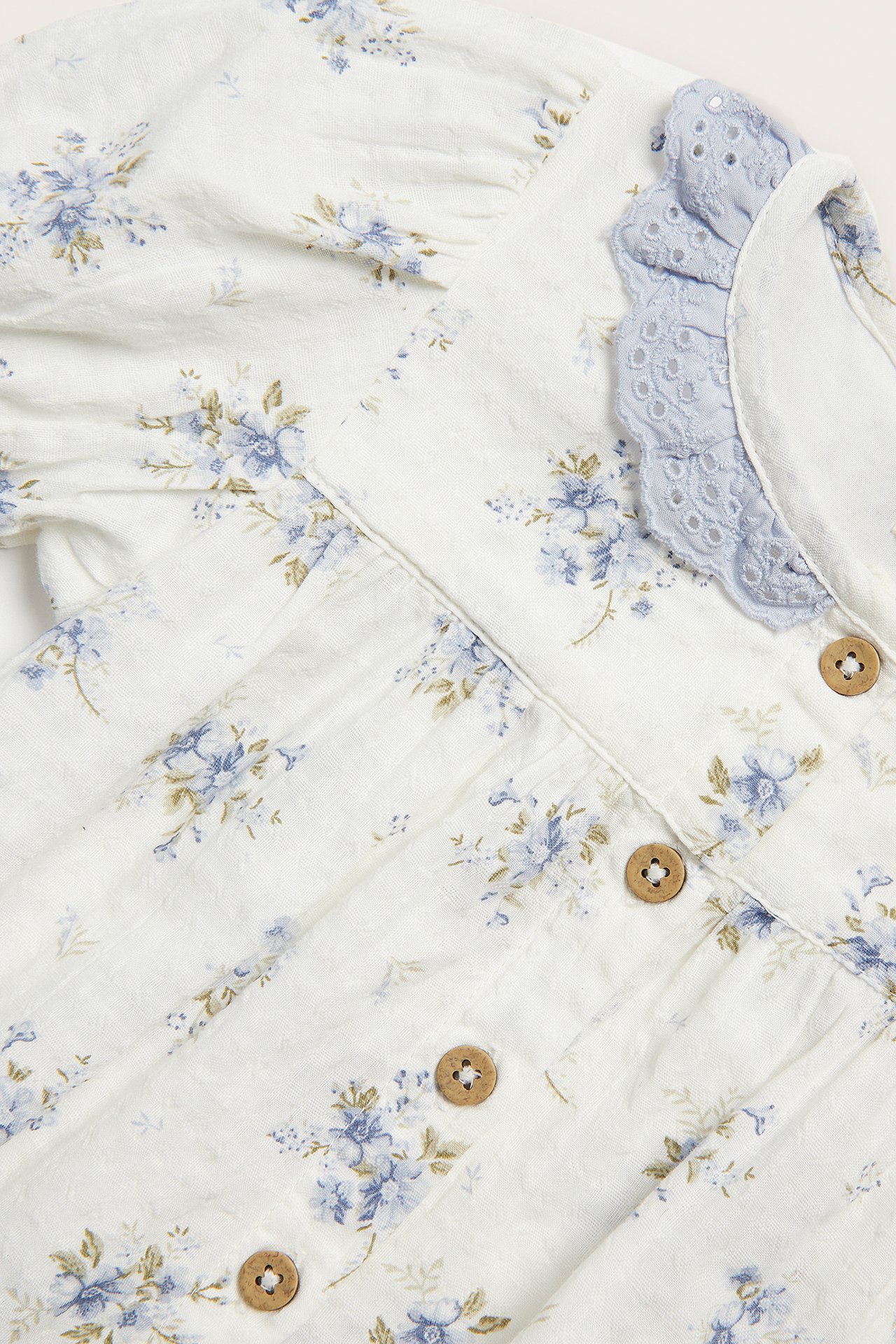 Close-up of an off-white baby bodysuit with blue floral print, light blue lace ruffle collar, and wooden front buttons.
