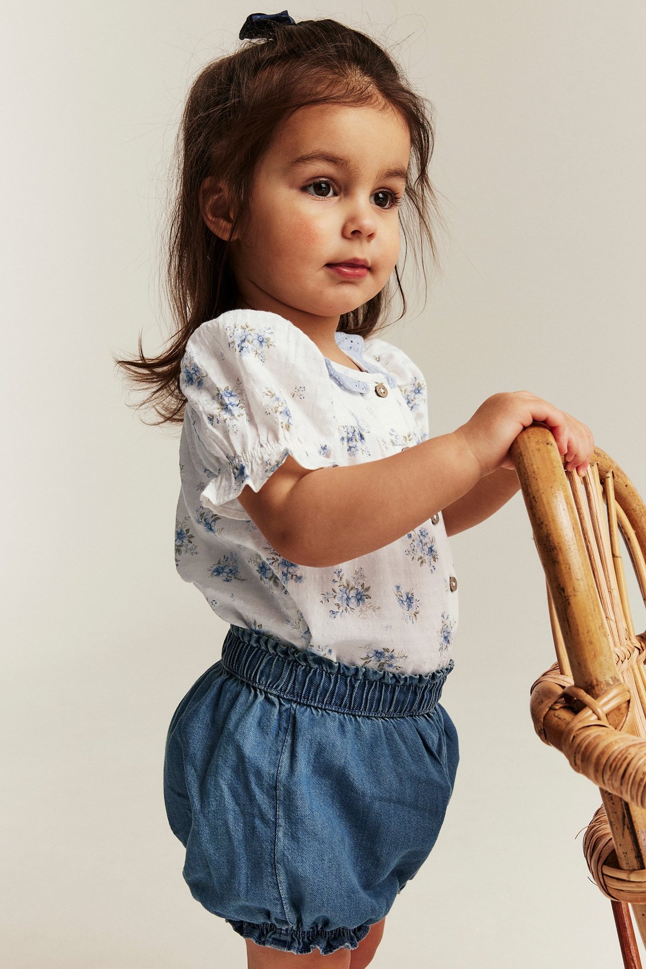 Babymädchen in offwhite geblümtem Kurzarm-Body und blauen Bloomer-Shorts, hält einen Rattan-Stuhl.