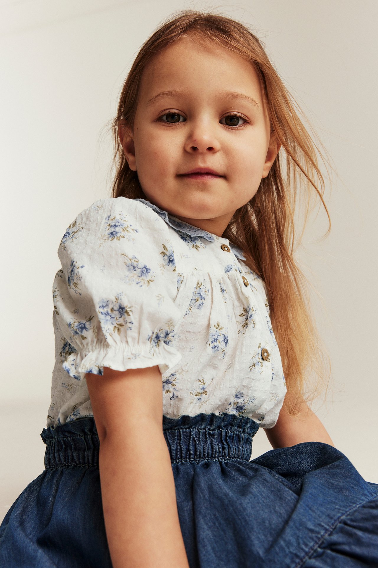 Kid wears offwhite cotton floral blouse with blue lace collar, front buttons, and a blue denim skirt.