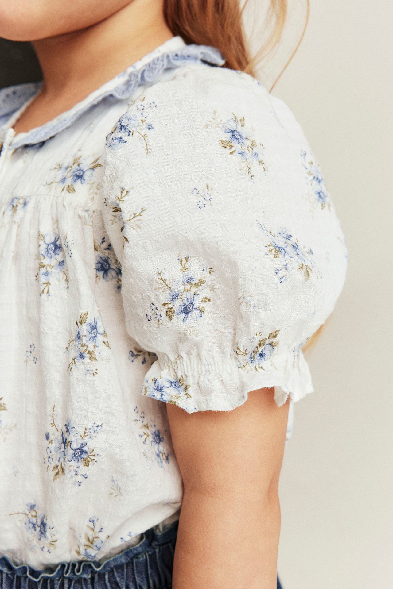 Kid's off-white floral blouse with blue flowers, puff sleeves, and light blue ruffled collar, close-up.