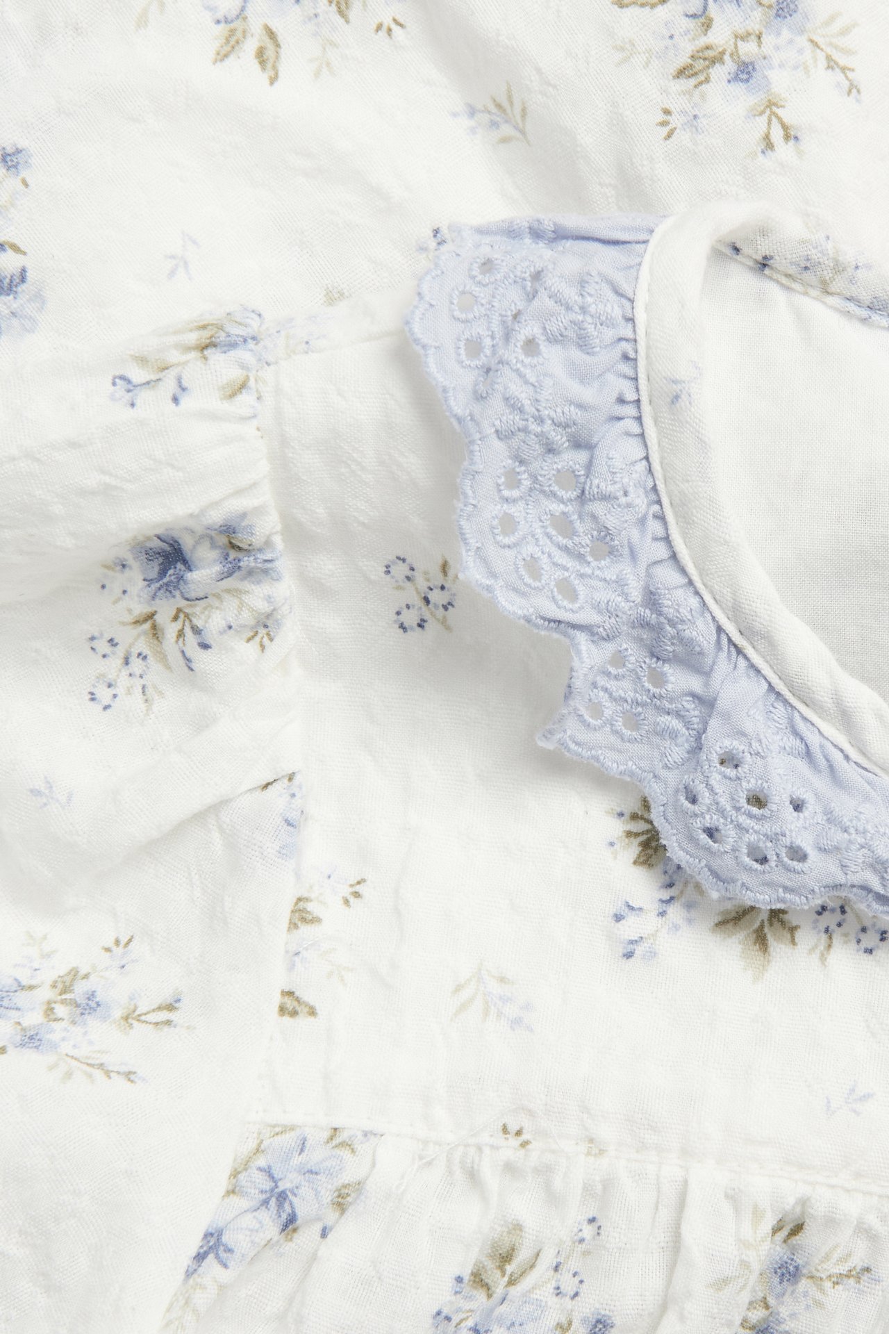 Close-up of an off-white woven blouse with blue floral pattern and a light blue lace ruffled neckline.