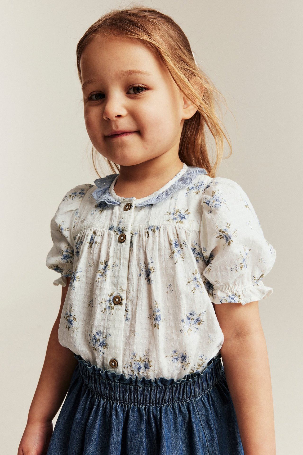 Front view of a kid in an off-white floral short-sleeved blouse with a blue lace collar and denim skirt.