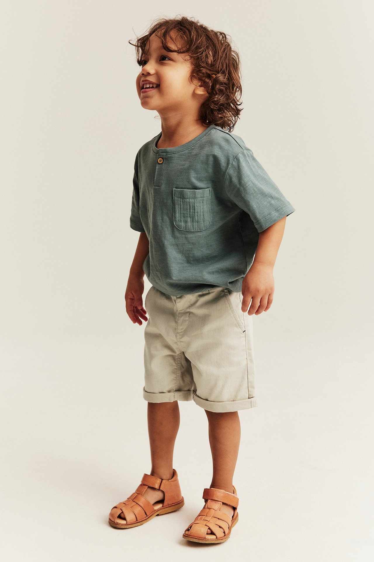 Side view of a kid wearing beige woven shorts with rolled-up legs, a green t-shirt, and brown sandals.