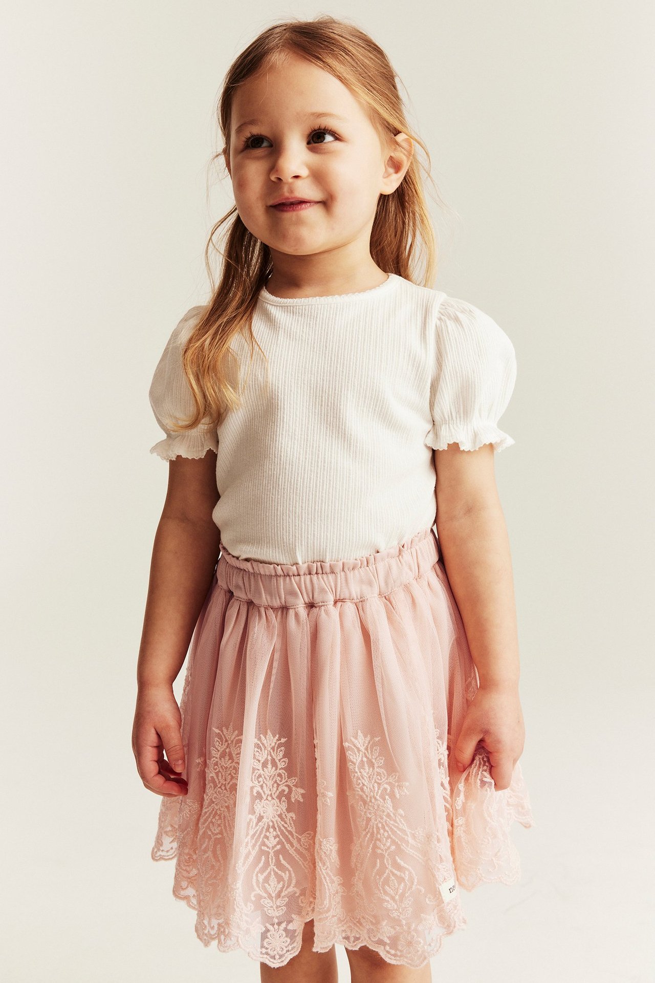 Front view of a kid wearing a light pink embroidered mesh skirt and a white ribbed top.