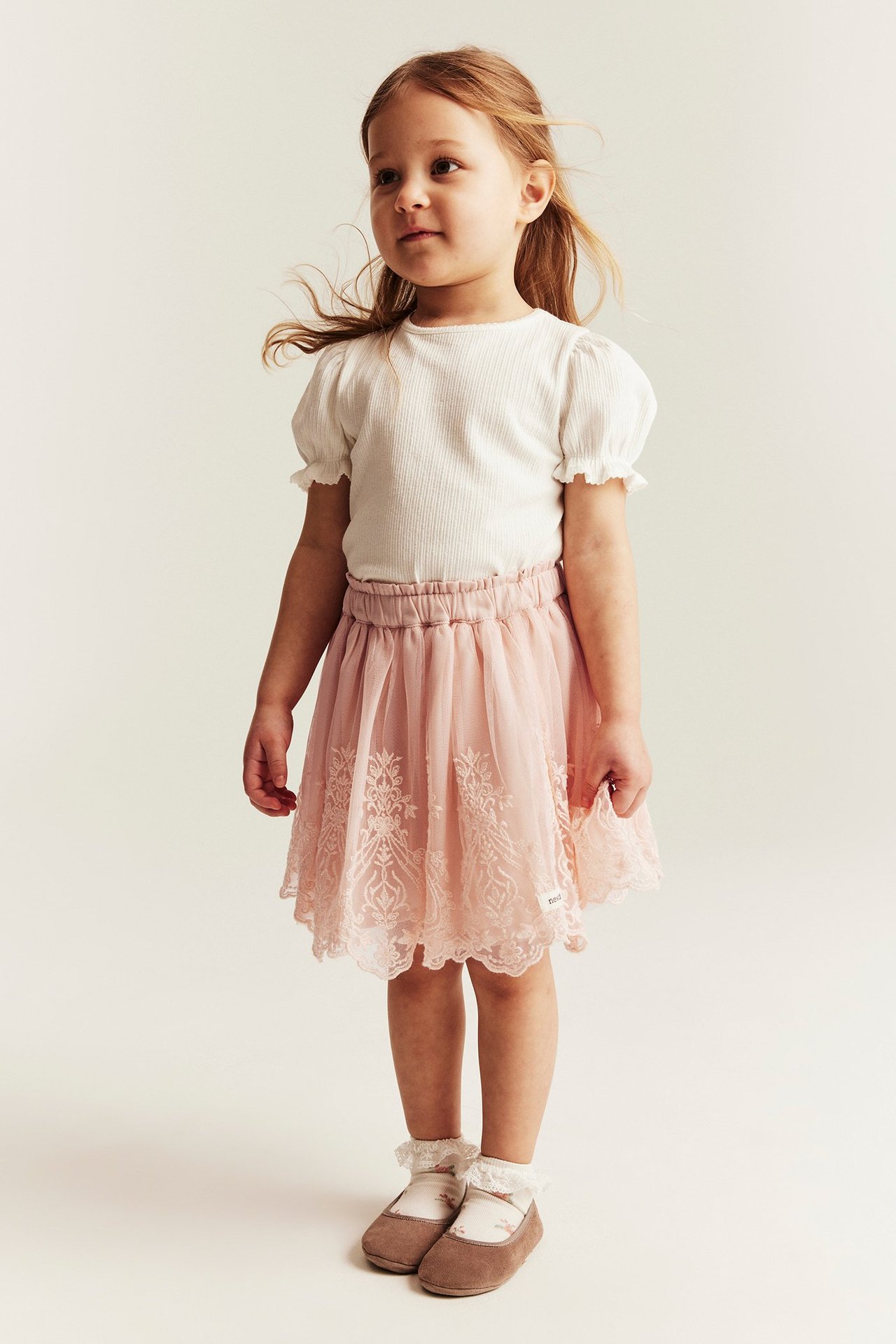 Front view of a kid in a light pink embroidered mesh skirt, white ribbed top, patterned socks, brown shoes.