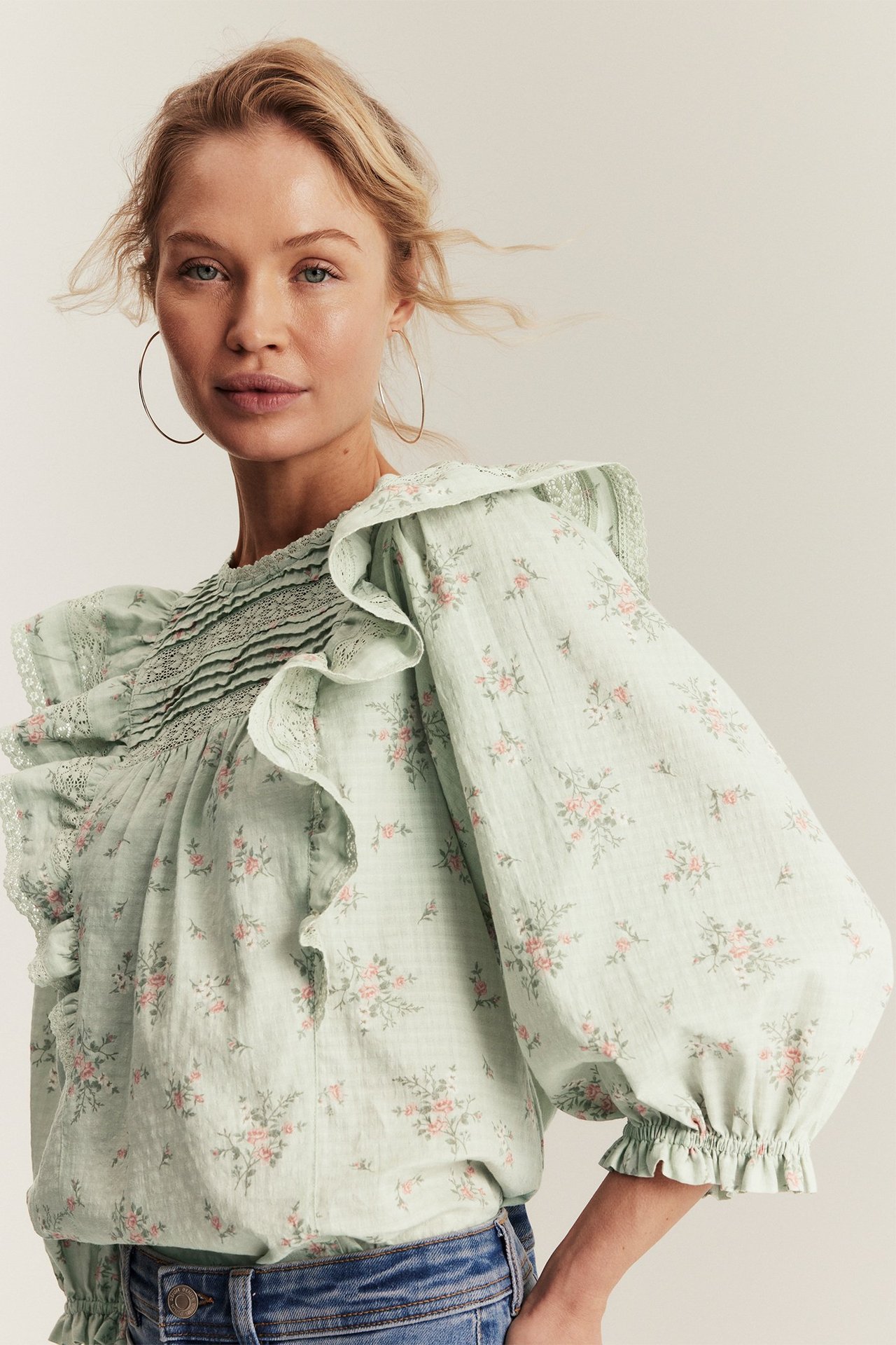 Vorderansicht einer Frau in hellgrüner floraler Baumwollbluse mit Rüschen, Puffärmeln, Jeans und Creolen.