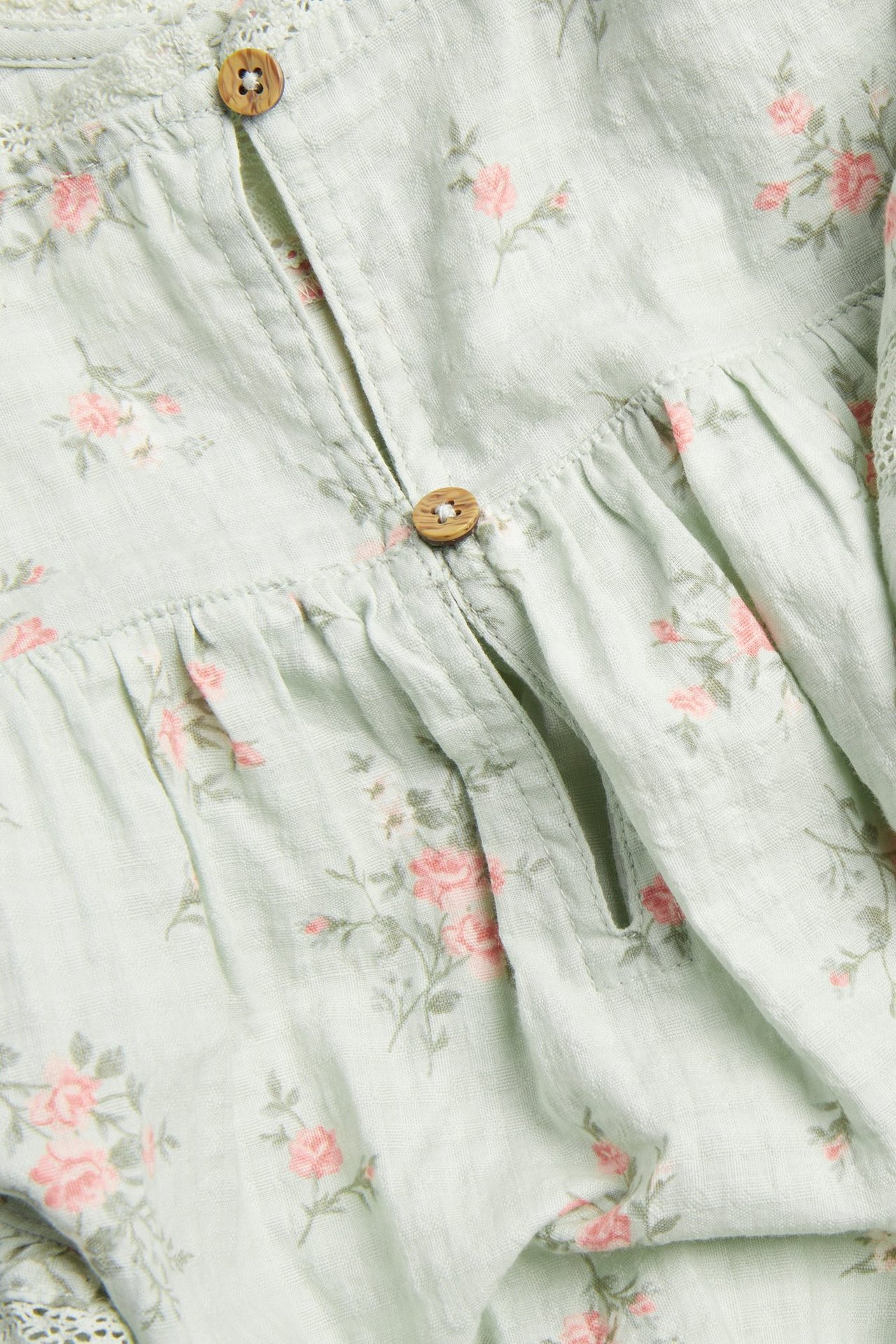 Close-up front view of a light green floral cotton blouse with pink roses, wooden buttons, and lace.