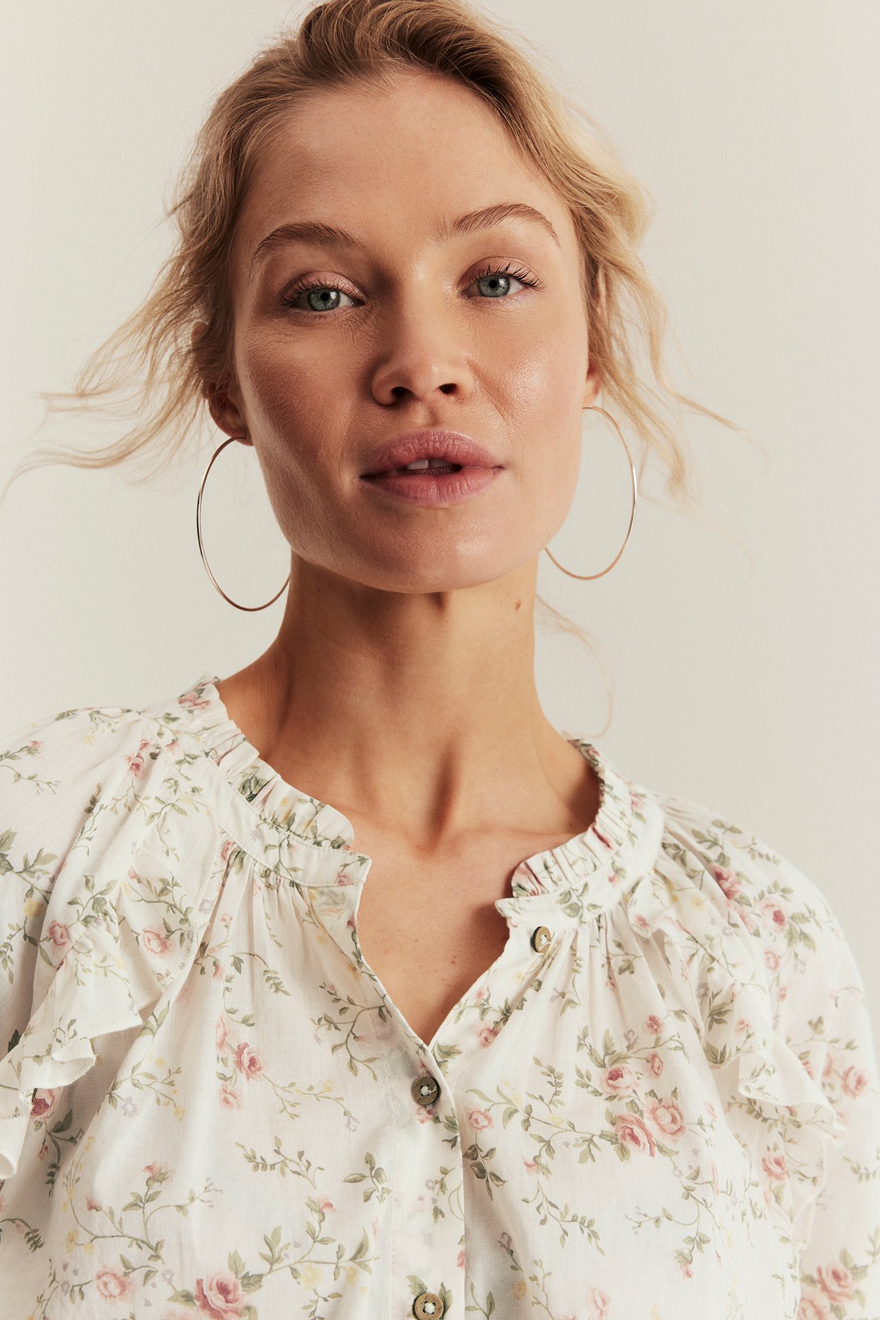 Woman wears off-white floral cotton blouse with ruffles, front buttons, and large hoop earrings.