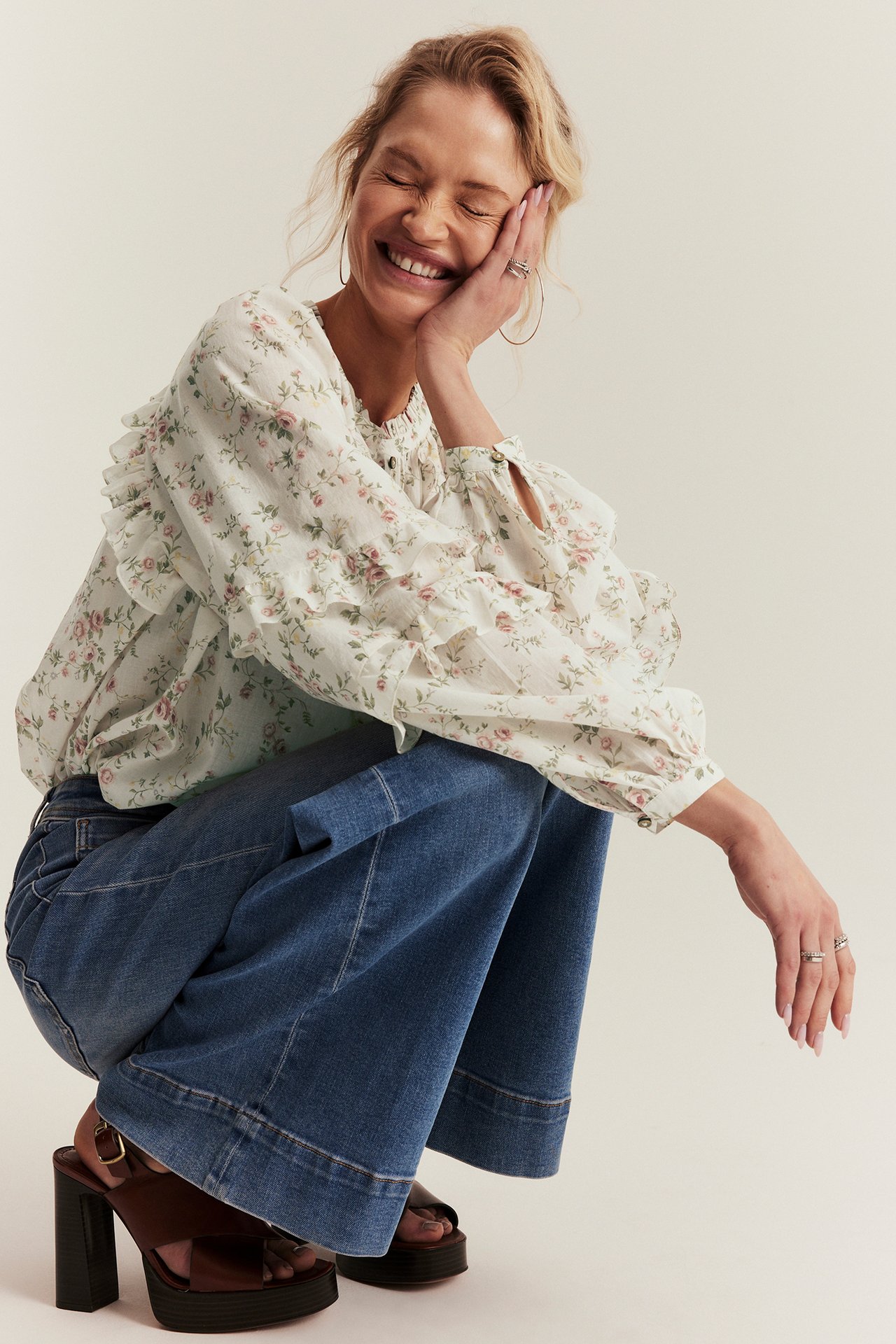 Smiling woman in an off-white floral ruffled cotton blouse, blue wide-leg jeans, and brown platform sandals, front-side view.