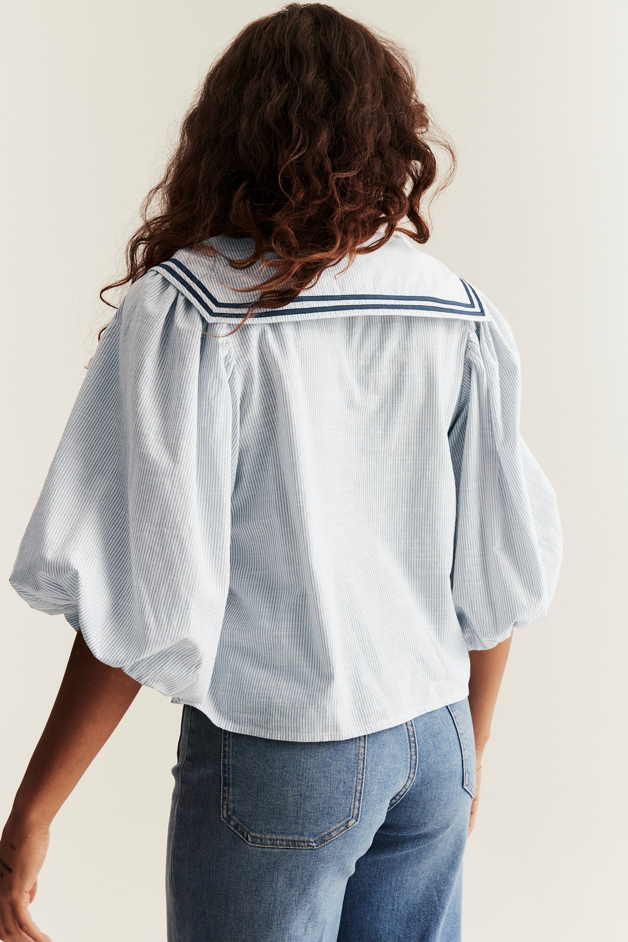Back view of woman in a light blue striped blouse with sailor collar, puff sleeves, and blue jeans.