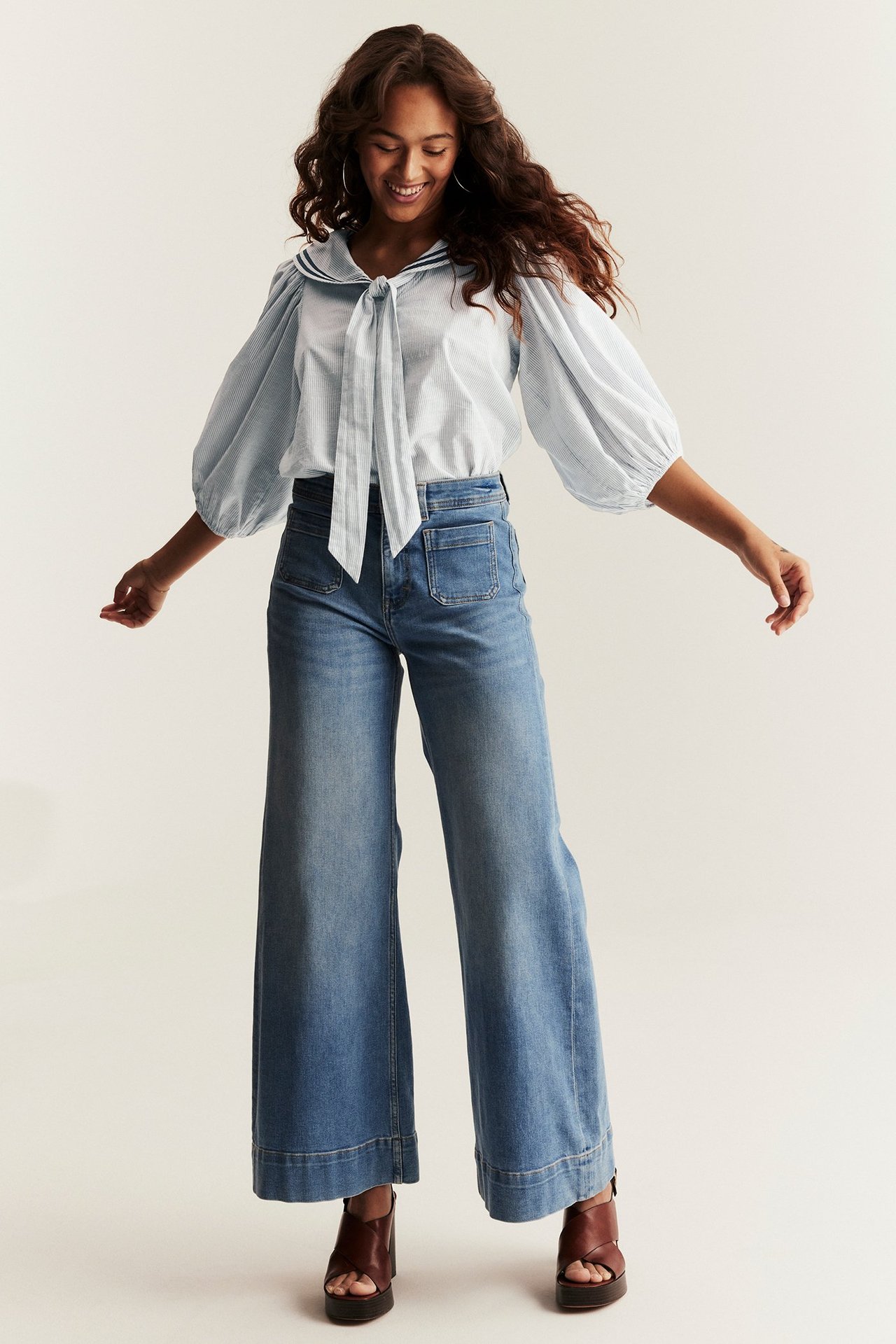 Front view: woman in light blue striped sailor blouse with tied bow, wide-leg jeans, brown sandals.